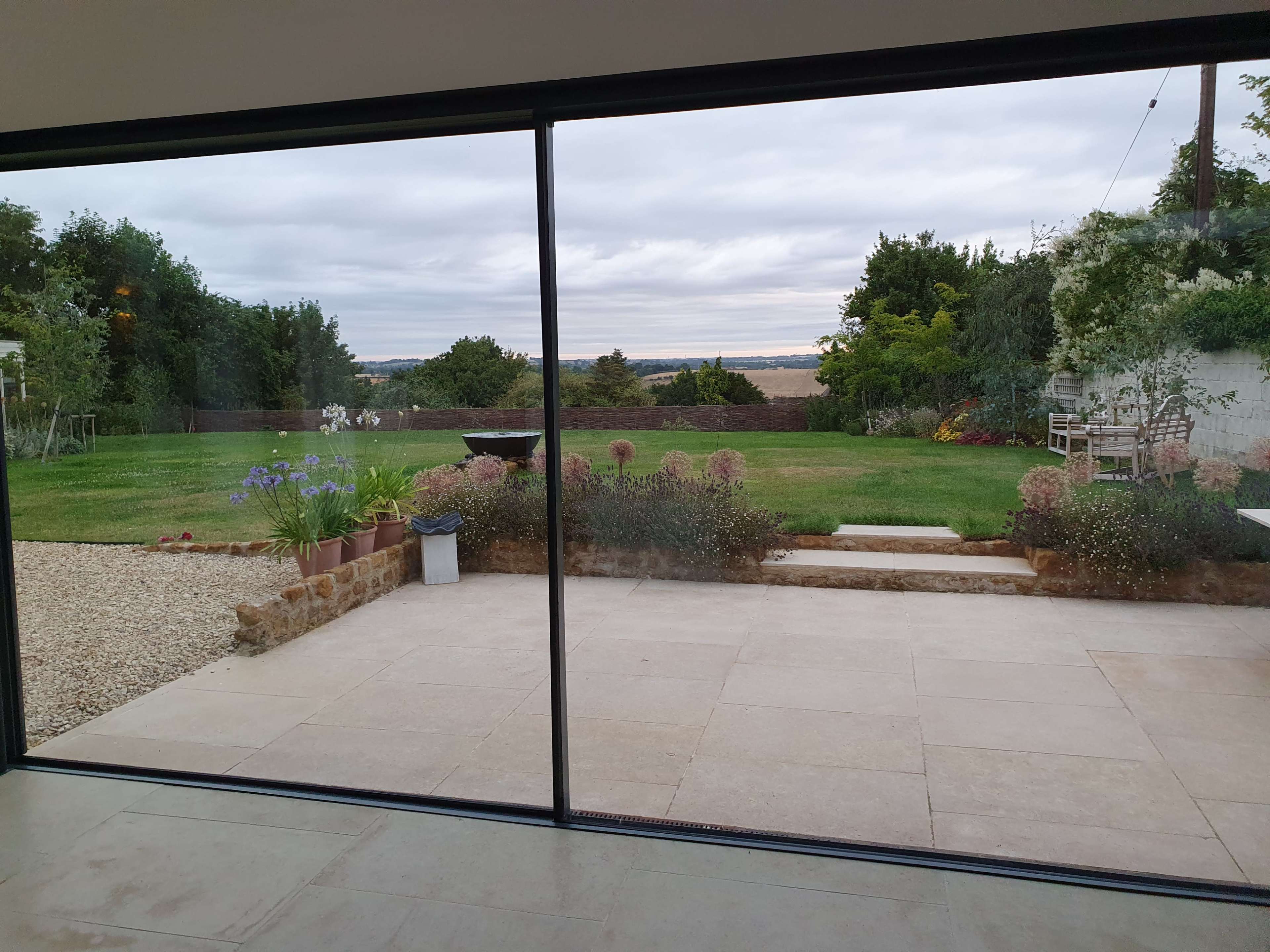 The image shows a landscaped garden with a grassy area and a view of fields, seen through large glass doors.