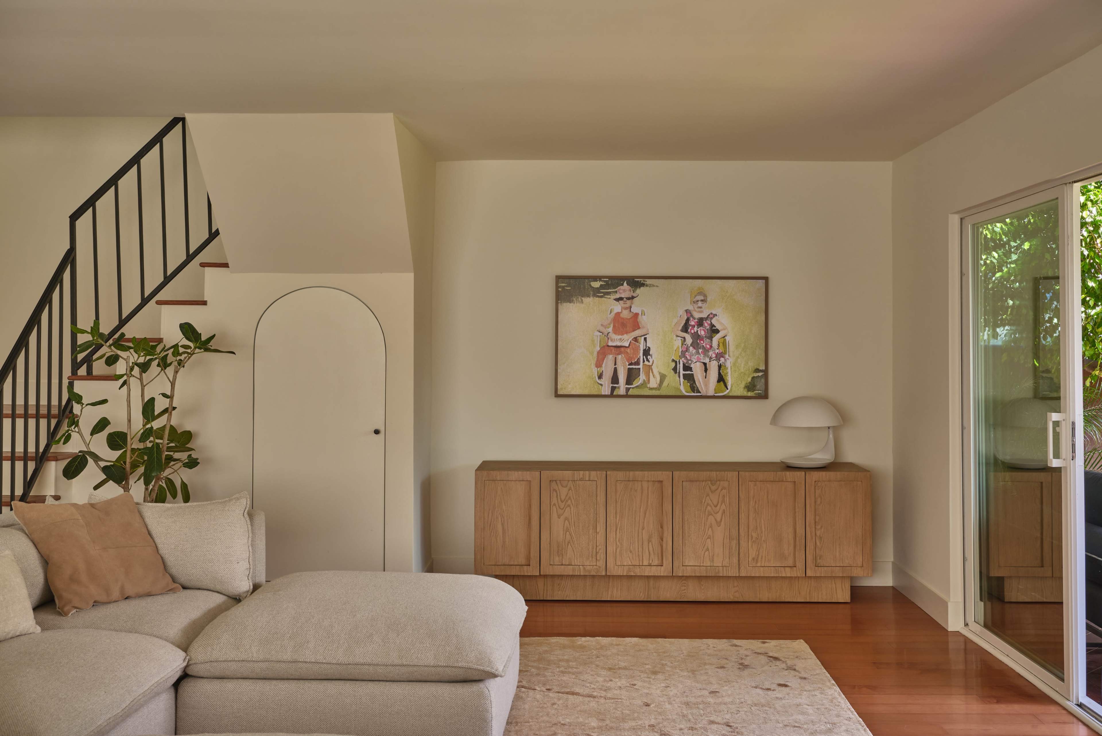 The image shows a modern living room featuring a light-colored sofa, a wooden sideboard, and a painting of two women on the wall.
