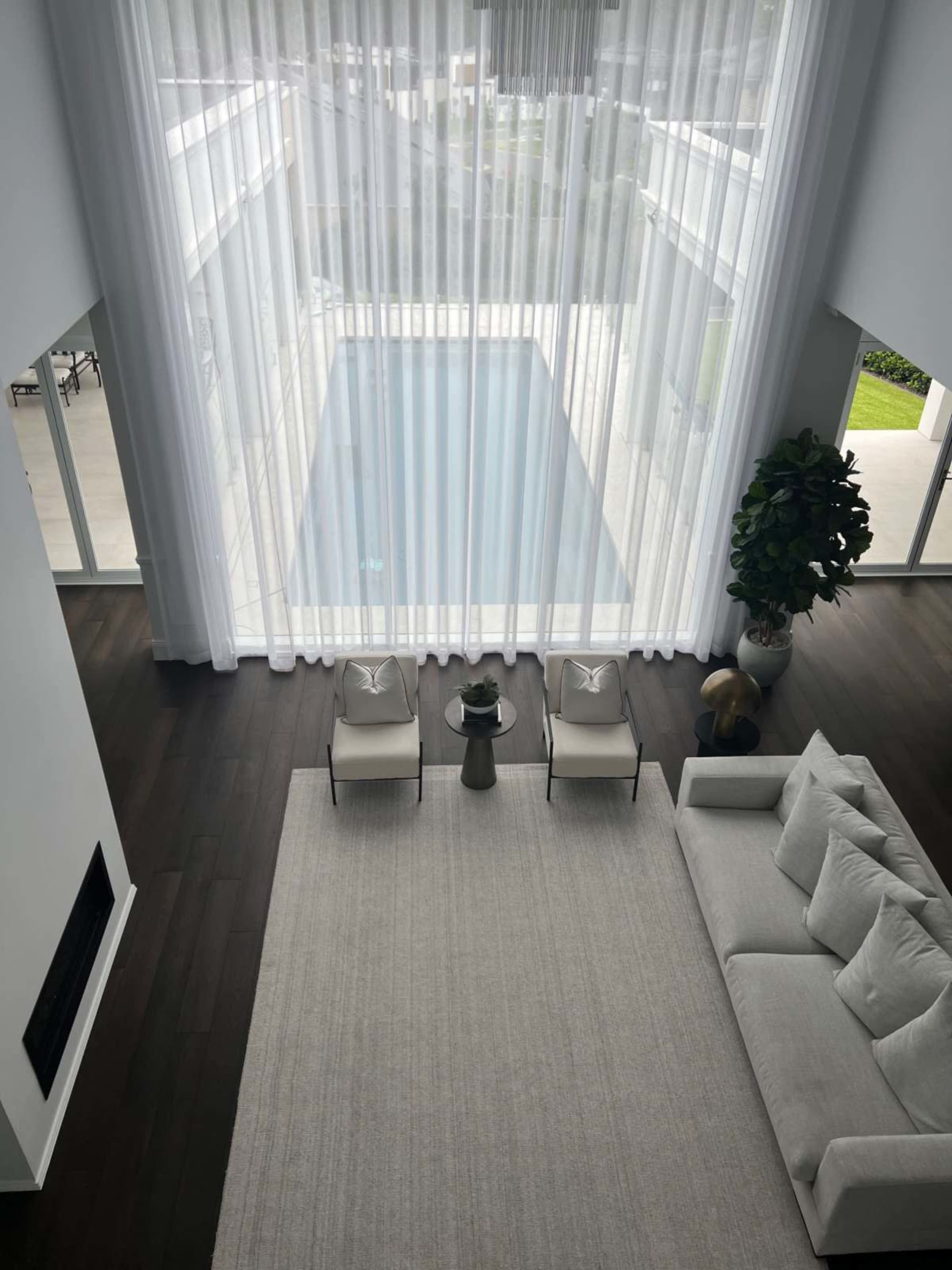 A spacious living area features a light-colored sofa and chairs, with large windows overlooking a pool area partially obscured by sheer curtains.