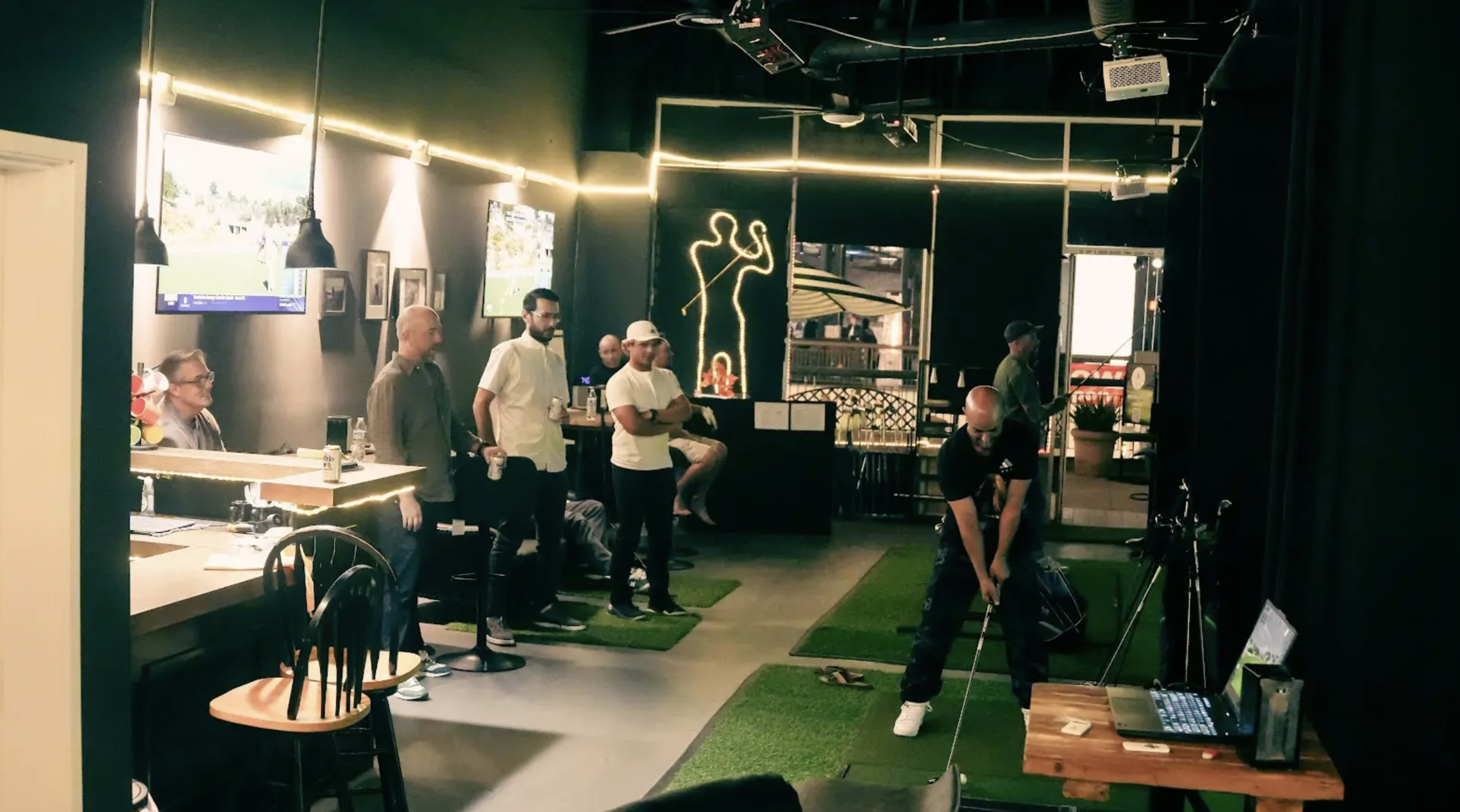 A group of men observe as one person practices a golf swing in an indoor golf simulation venue.