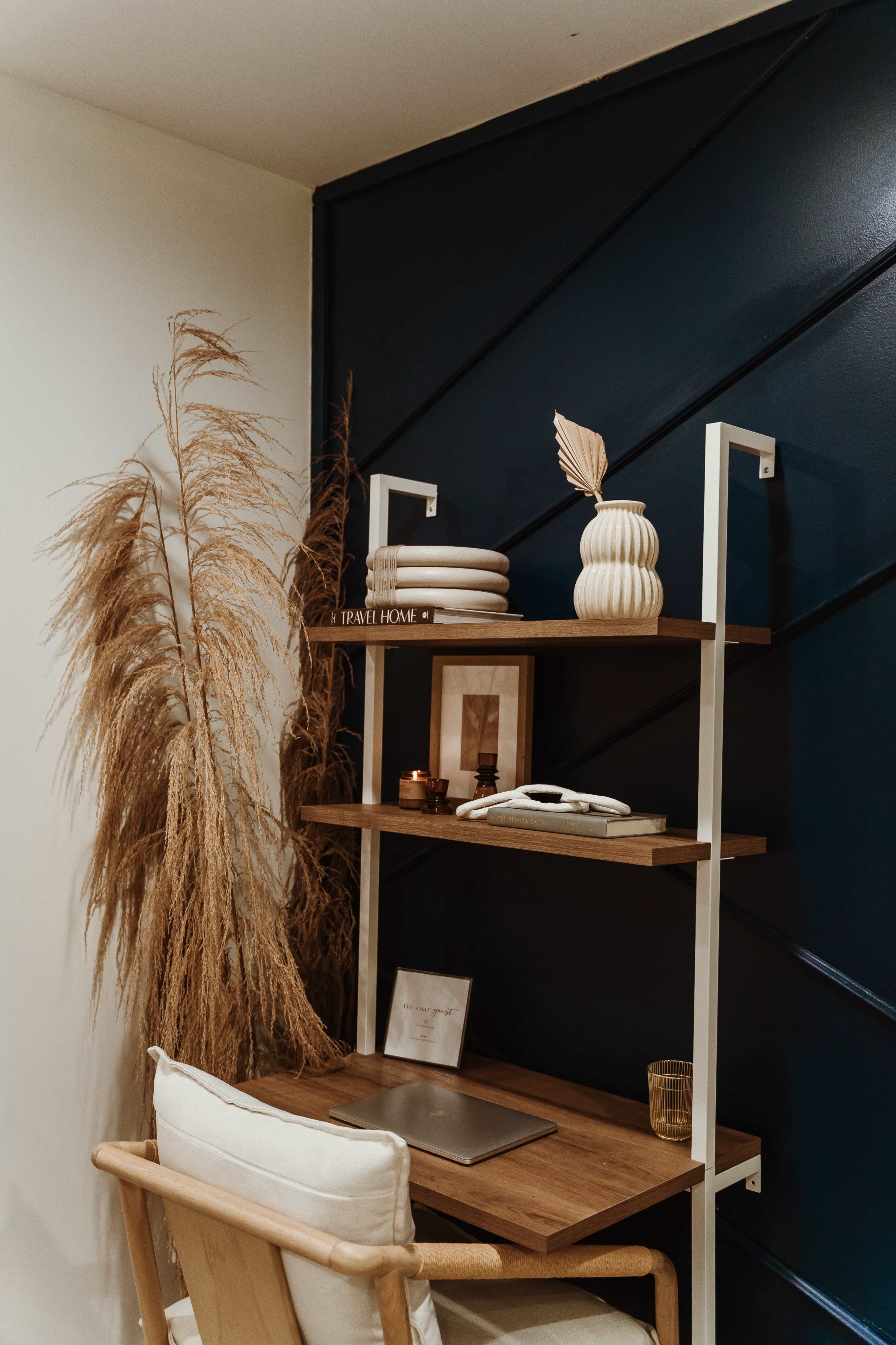 A modern workspace featuring a wooden desk with a laptop, a decorative shelf with vases and books, and a plant in the corner against a dark blue wall.