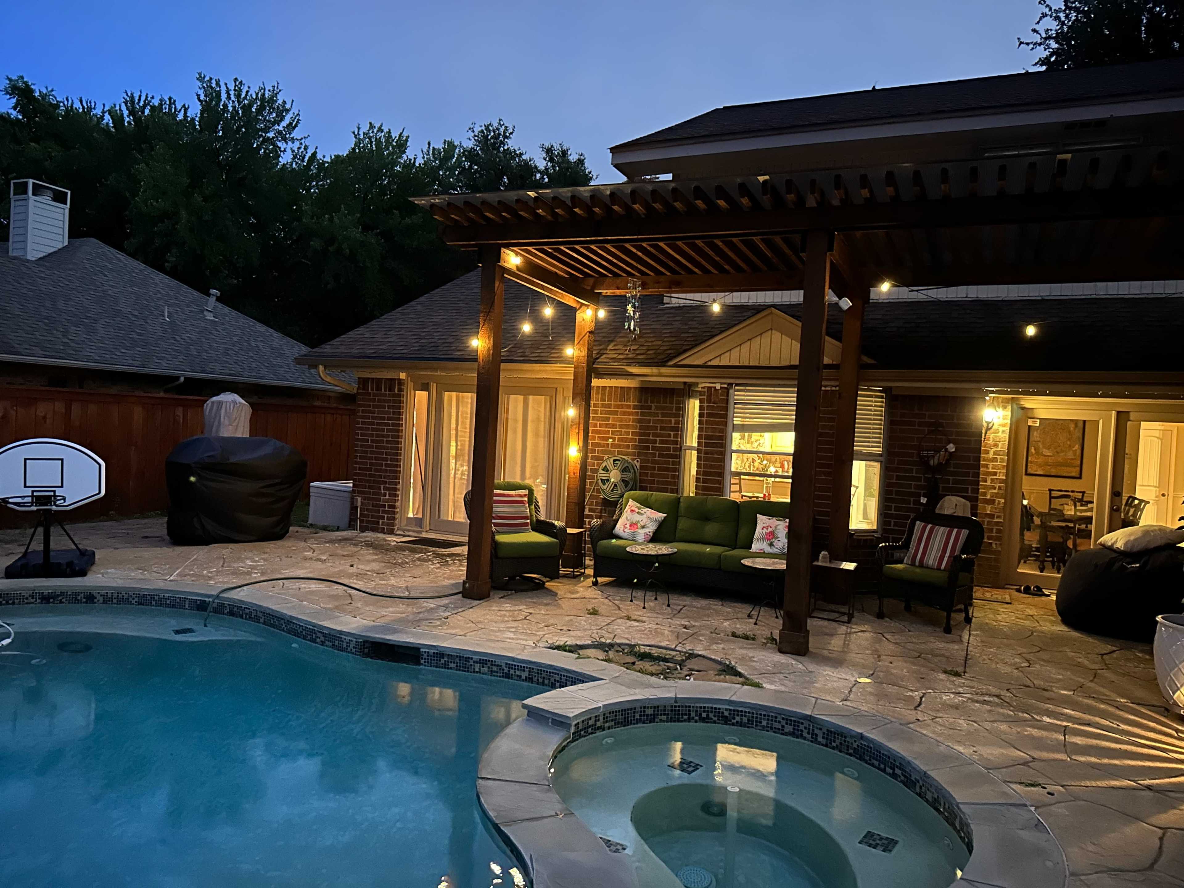 The image shows a backyard with a pool, hot tub, patio furniture, and lights strung along a wooden pergola at dusk.