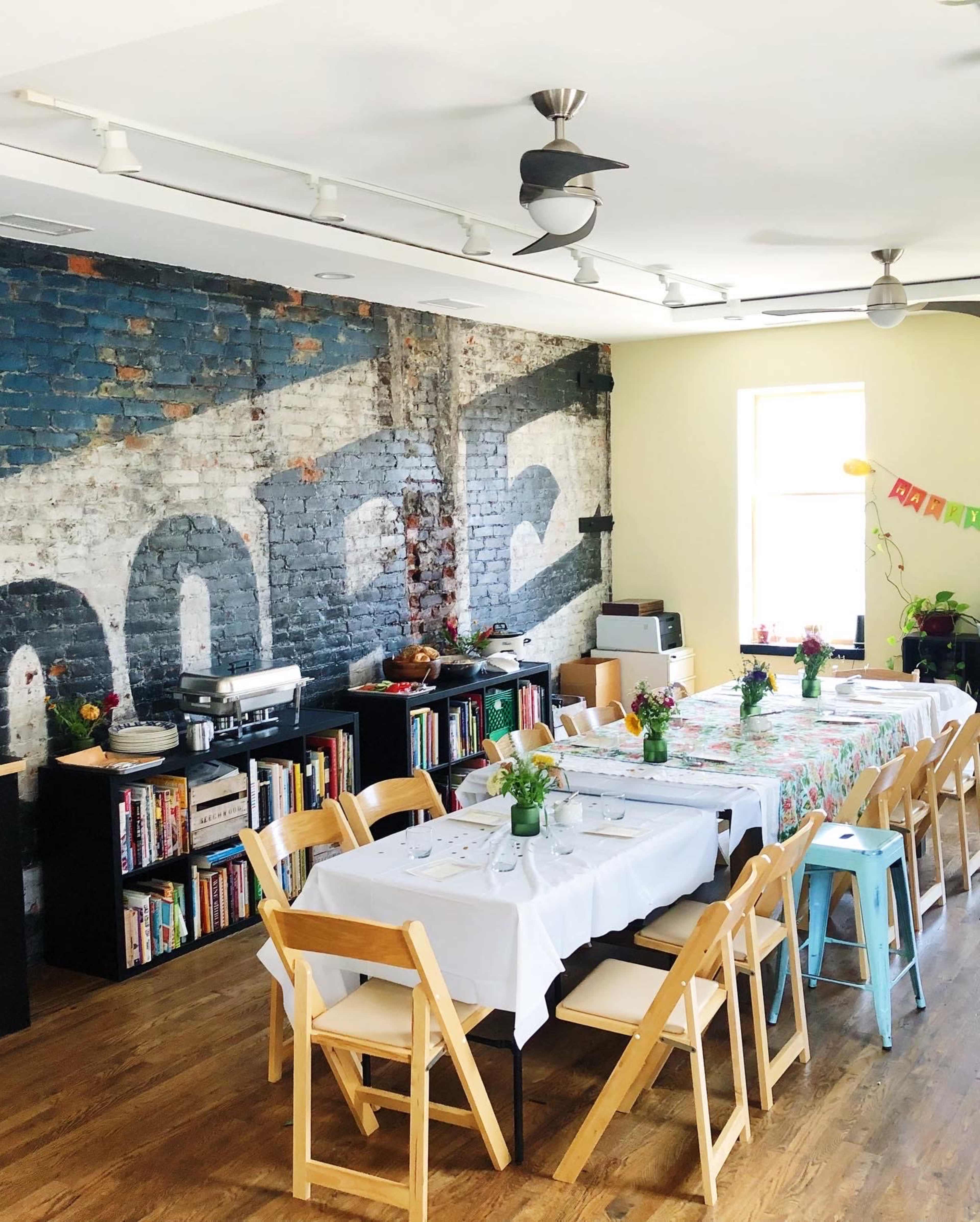 A dining area features multiple tables set with flowers, surrounded by bookshelves and a brick wall displaying a faded mural.