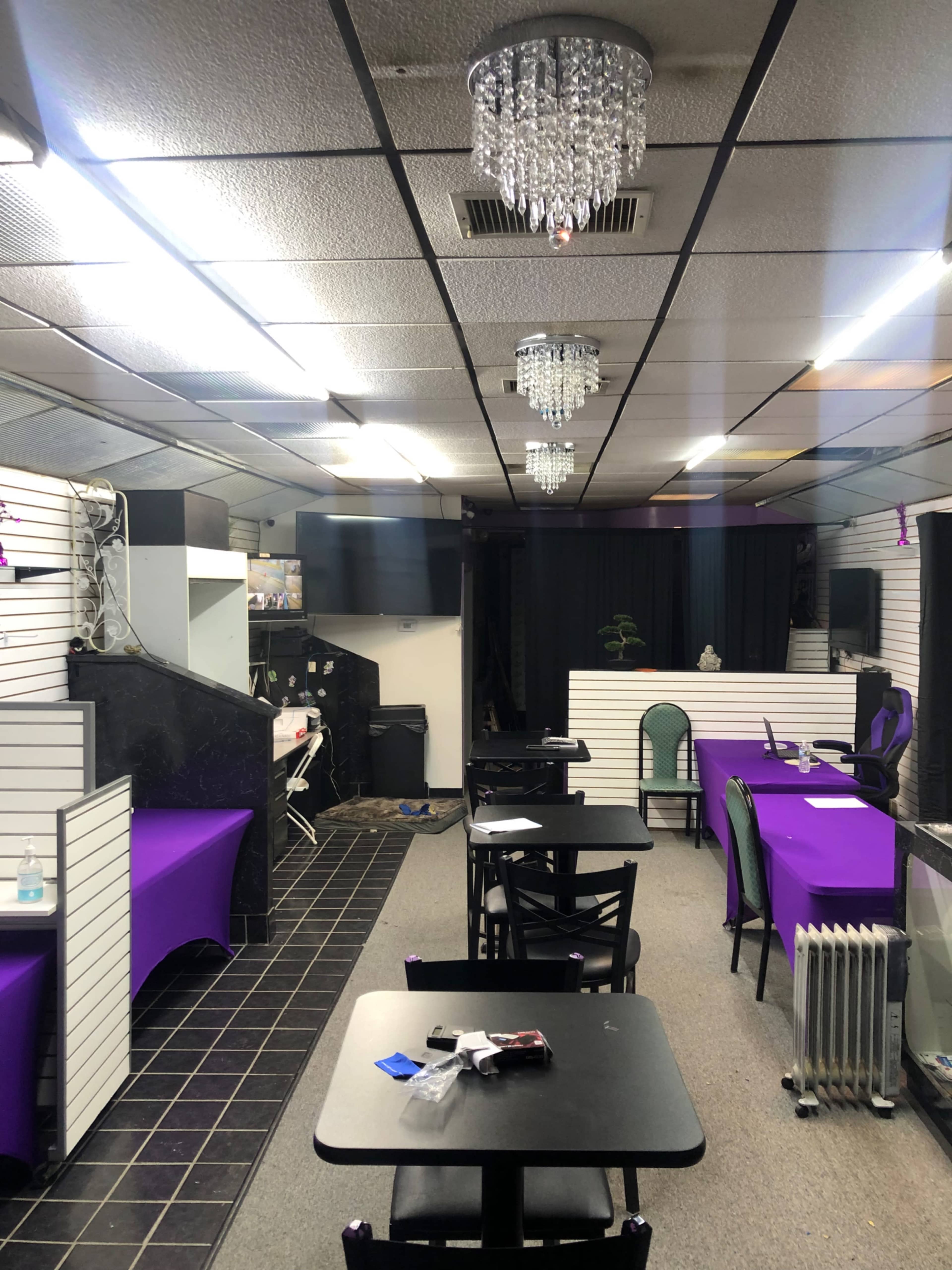 The image shows the interior of a small cafe or event space featuring empty tables with purple table covers, chandeliers, and a black curtain at the back.