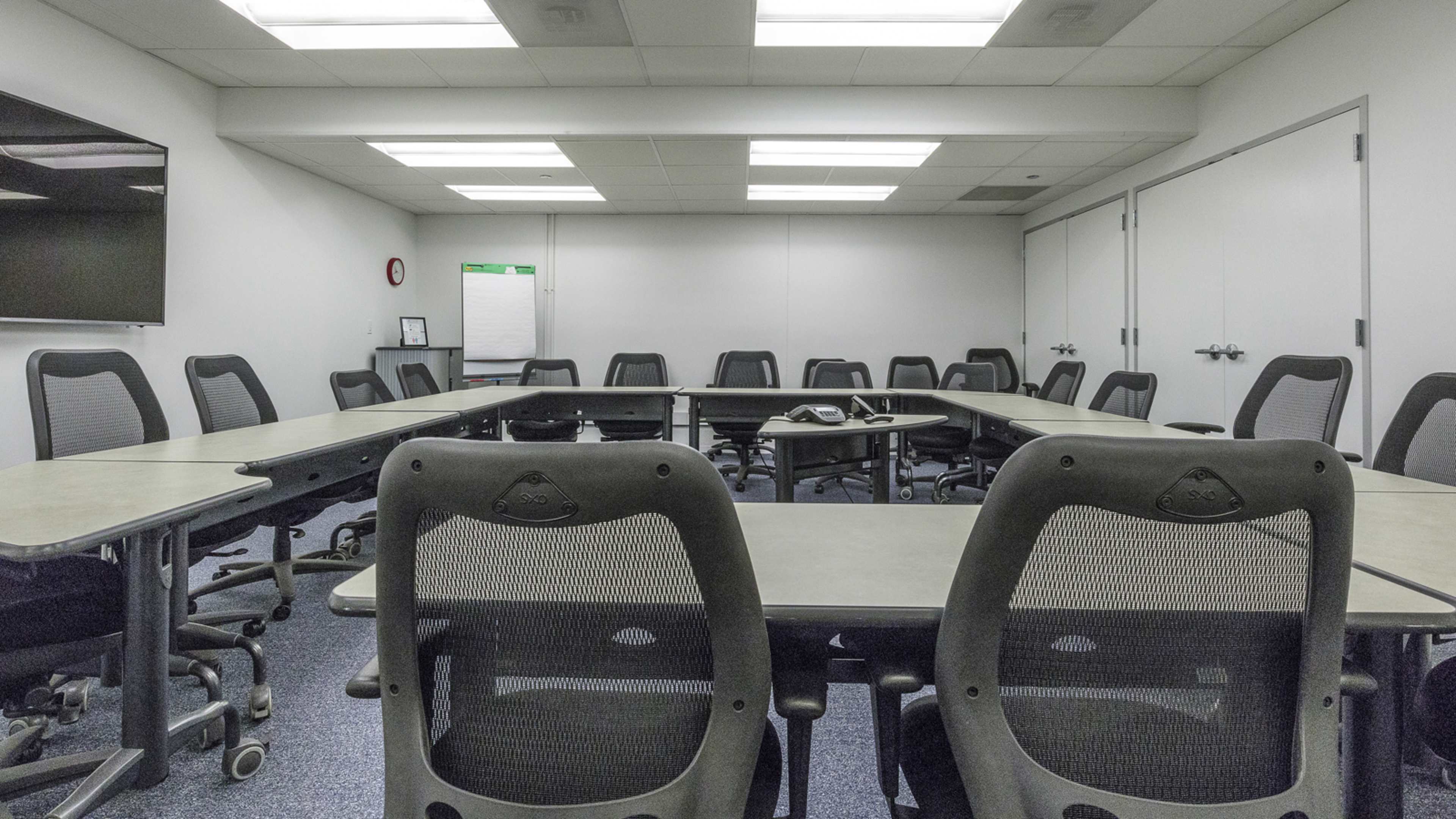 A conference room with a large oval table surrounded by black chairs and a wall-mounted screen at one end.