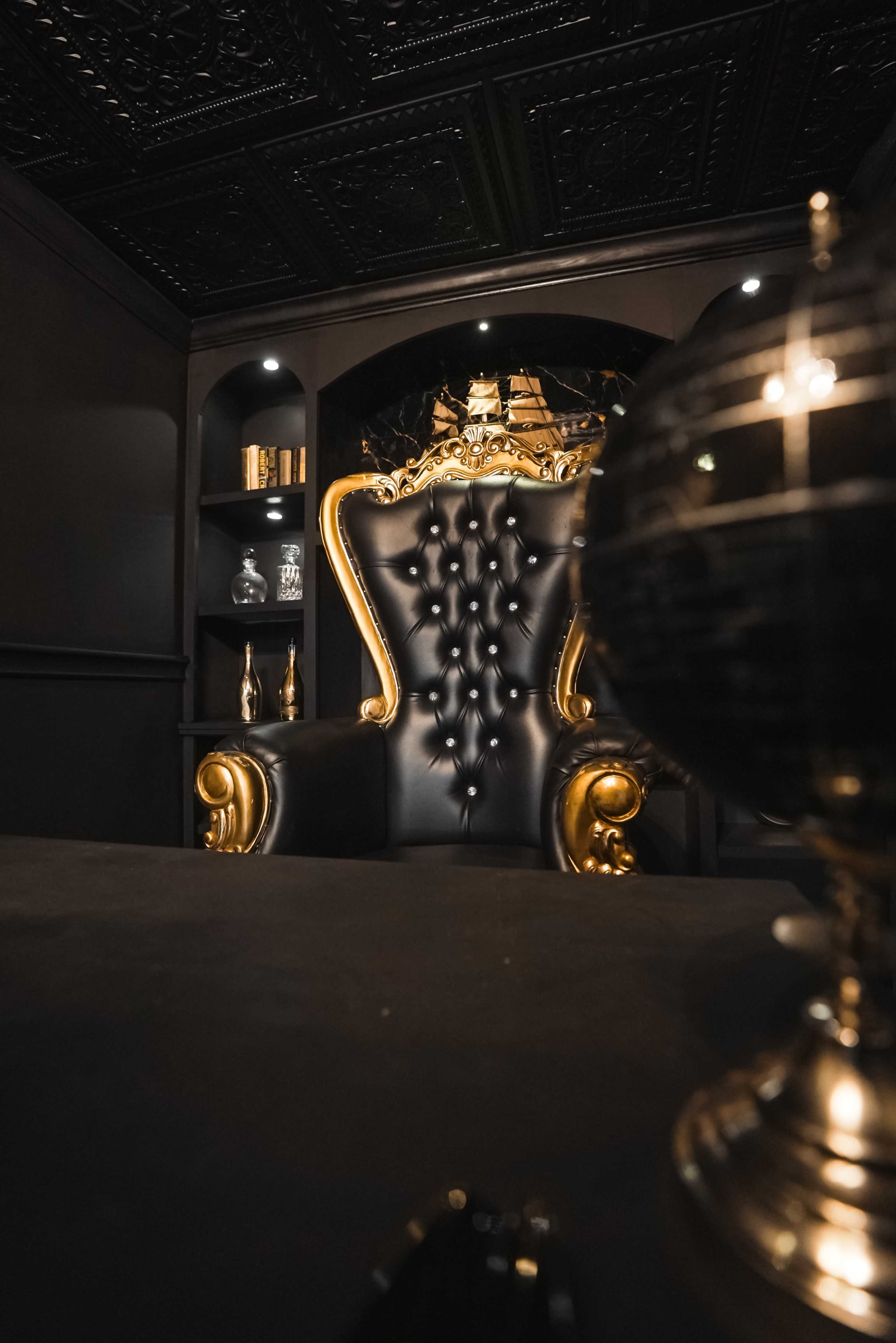The image shows a luxurious black and gold tufted armchair in a dark room with shelves displaying decorative items and a globe on a table.