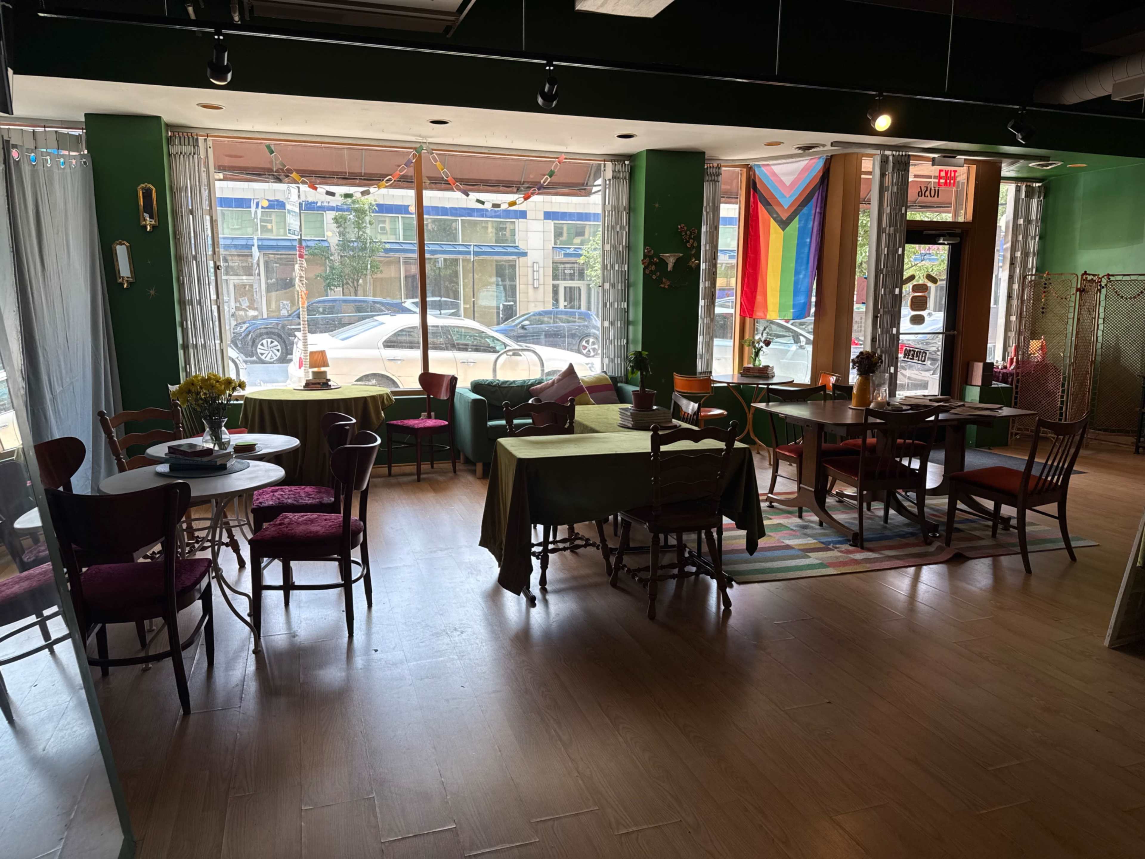 The image shows the interior of a café with various tables and chairs arranged around a spacious area, featuring large windows that allow natural light to enter.