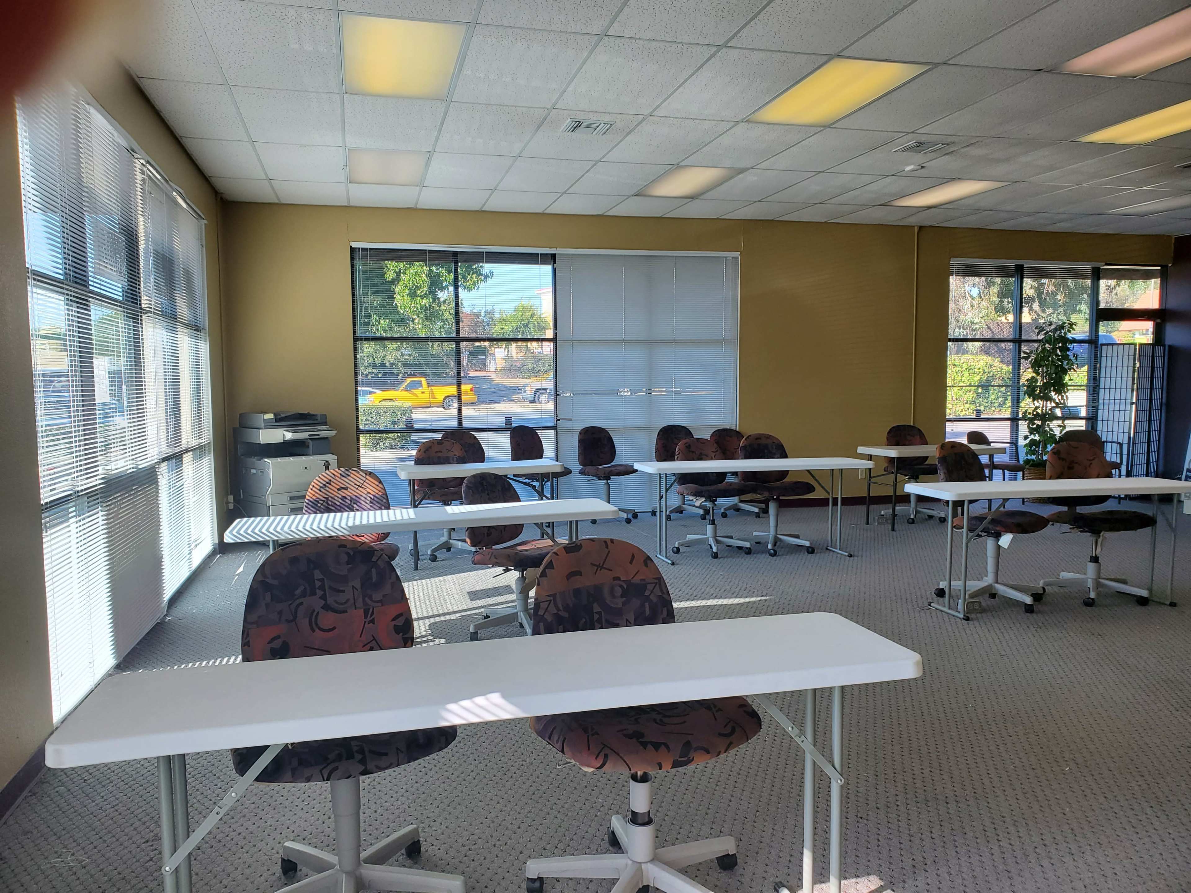 A brightly lit classroom with several white tables and patterned chairs arranged in a spacious layout, and large windows letting in natural light.