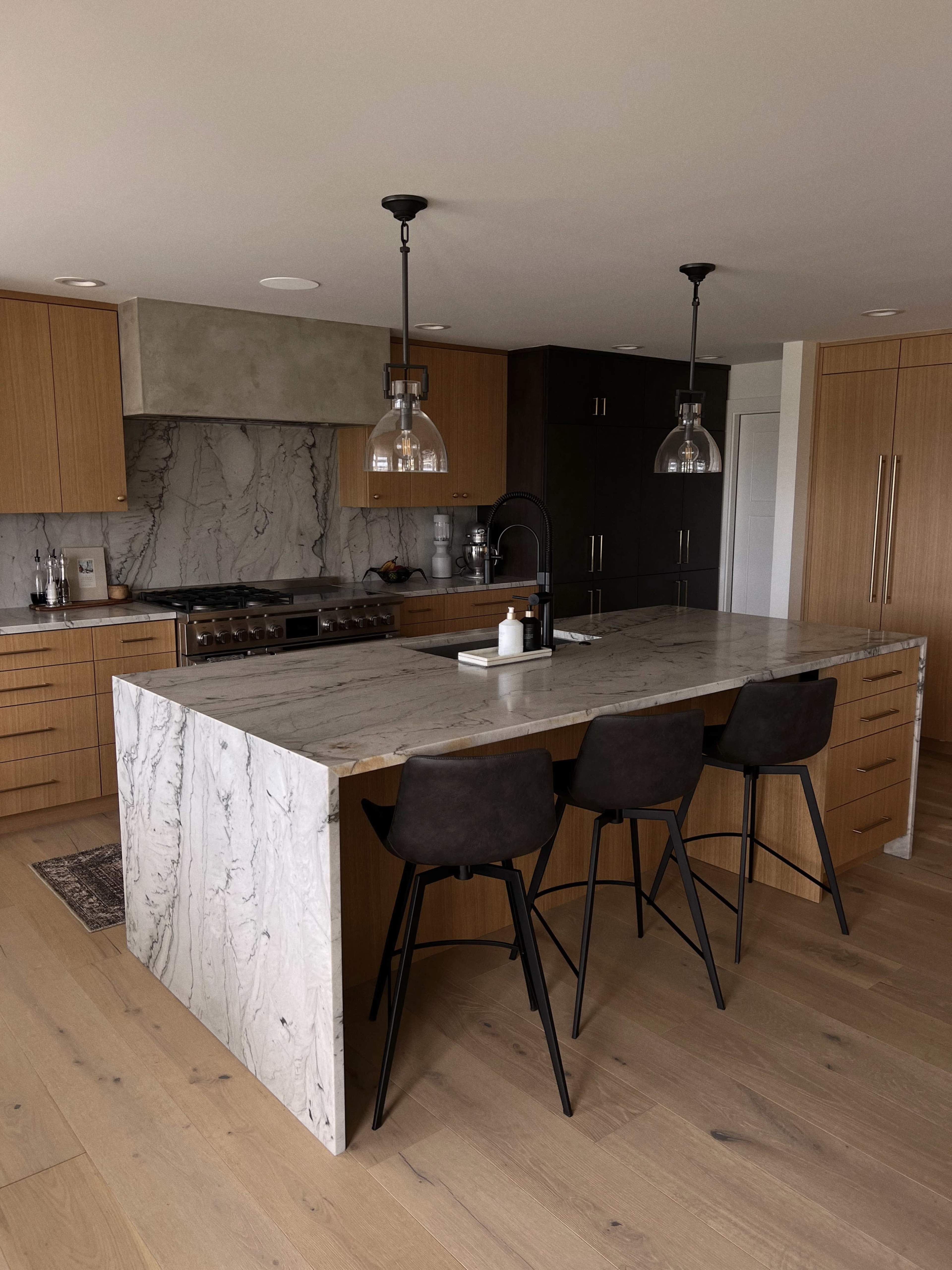 The image shows a modern kitchen featuring a large marble island with three black stools, wooden cabinetry, and stylish pendant lights.