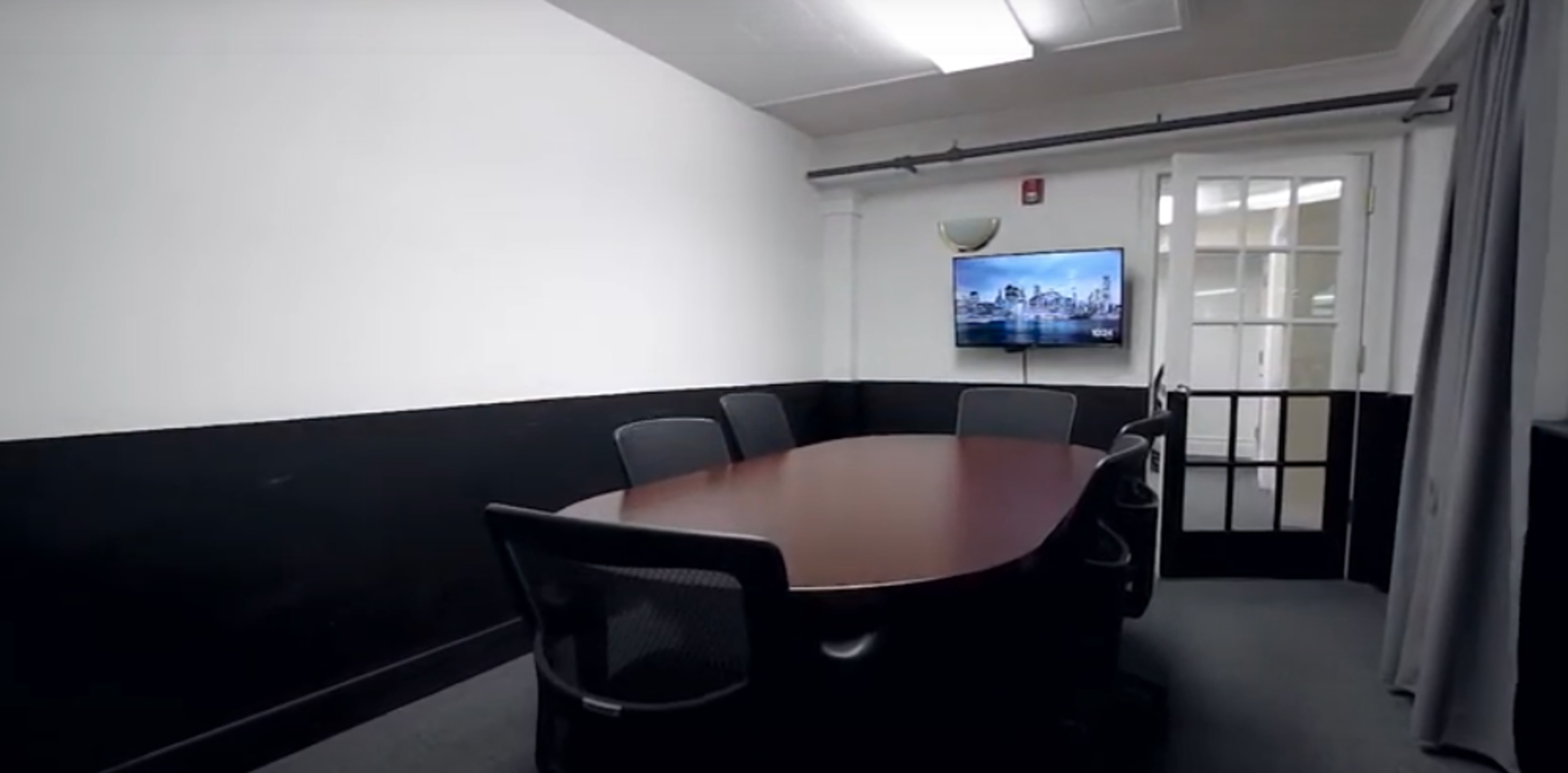 A conference room features a large oval table surrounded by chairs and a mounted television on the wall.