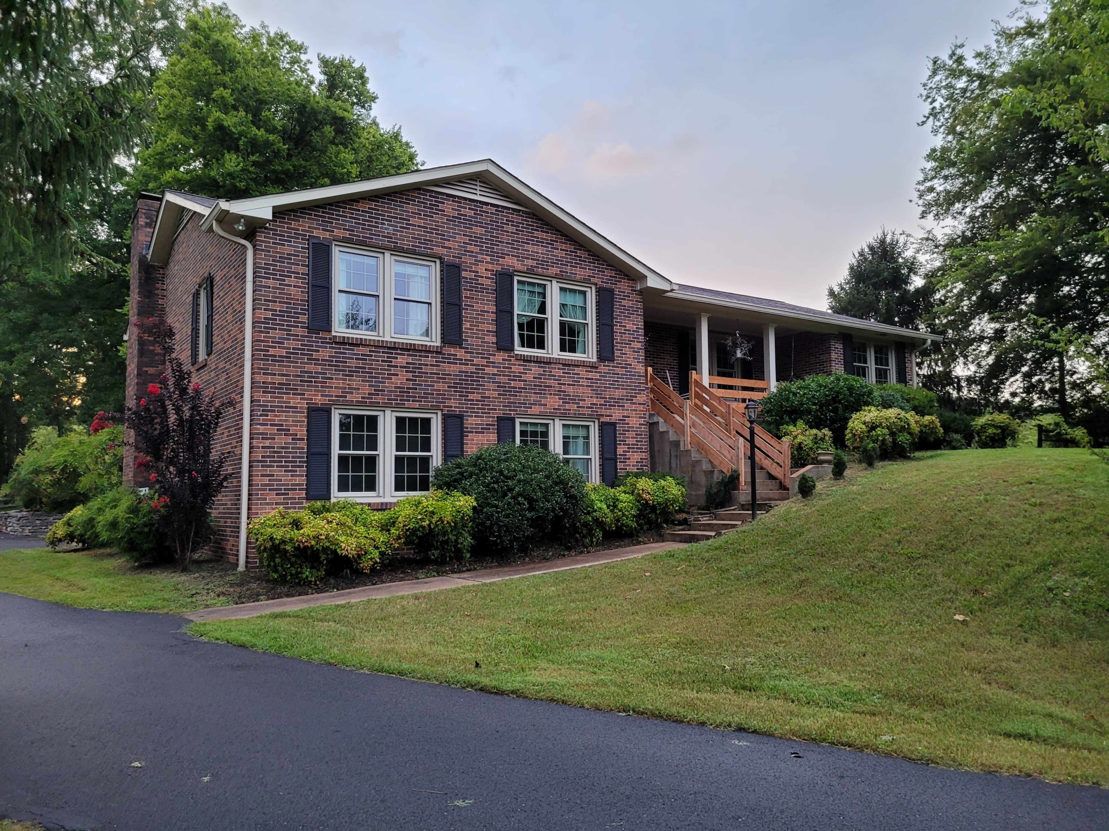 A two-story brick house with blue shutters sits on a grassy slope, accompanied by well-kept shrubbery and a gravel driveway.