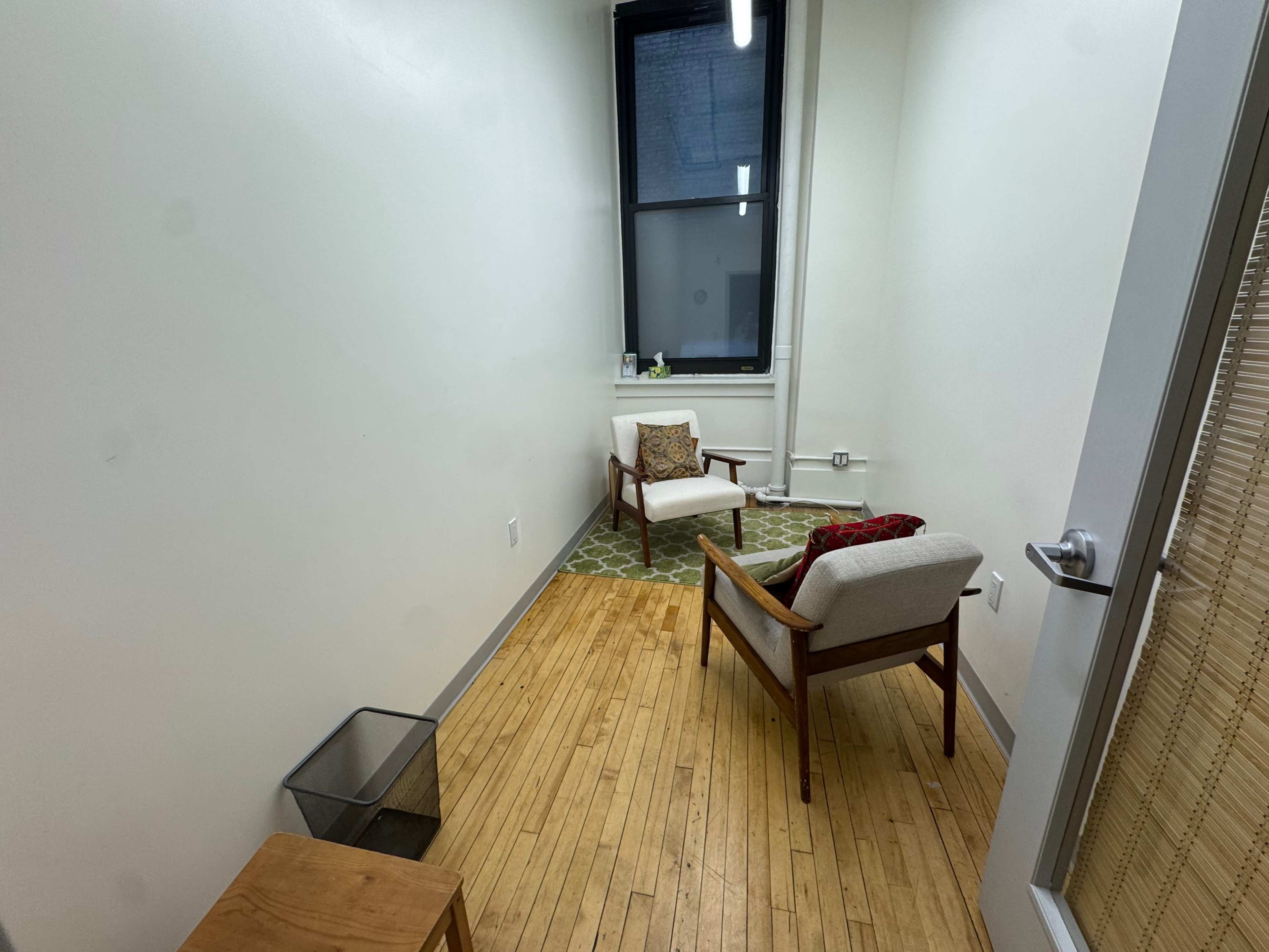 The image shows a small, sparsely furnished room with two chairs, a small table, a wastebasket, and a large window.