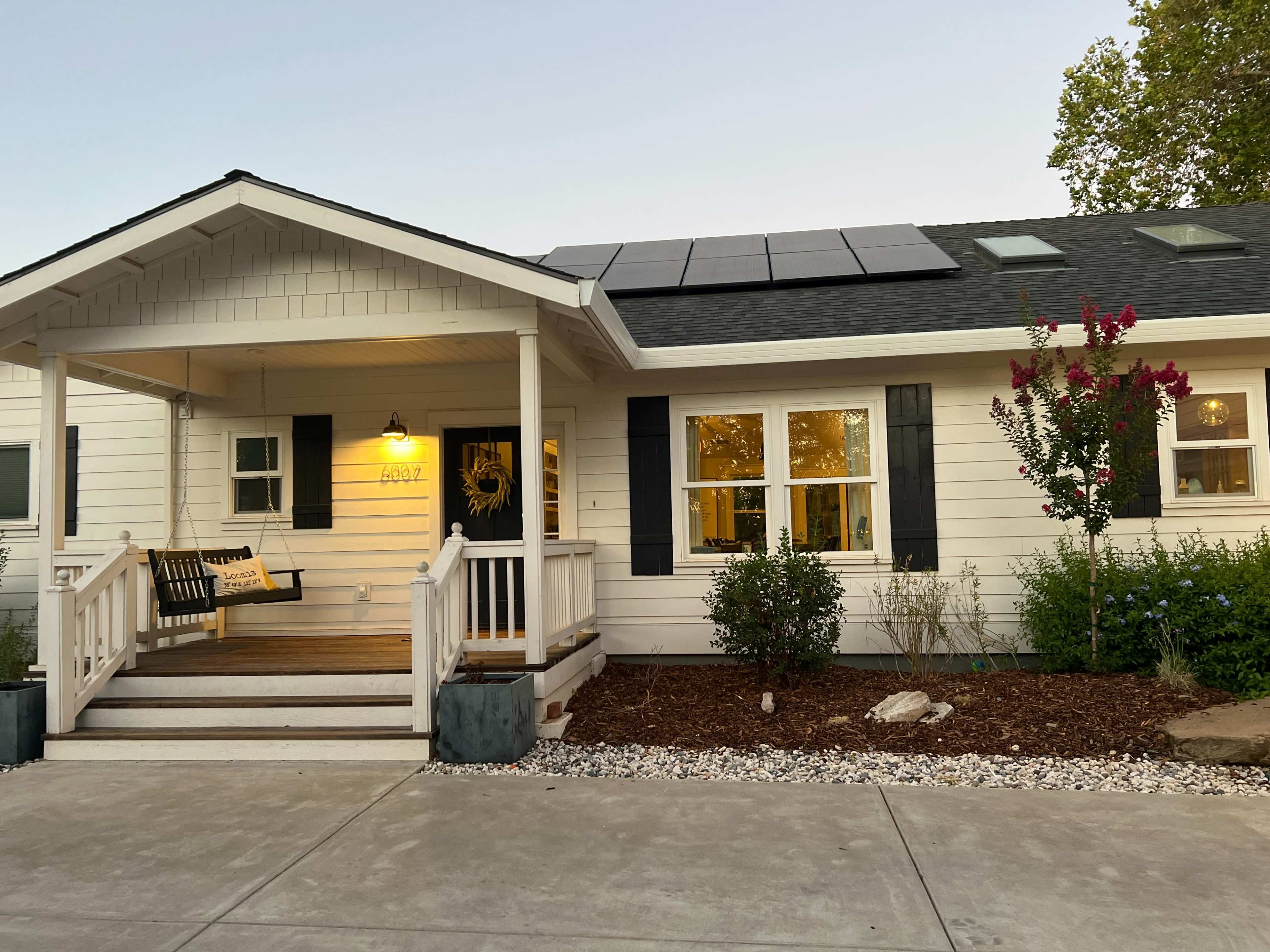 A single-story house features a front porch with a swing, solar panels on the roof, and landscaped gardens.