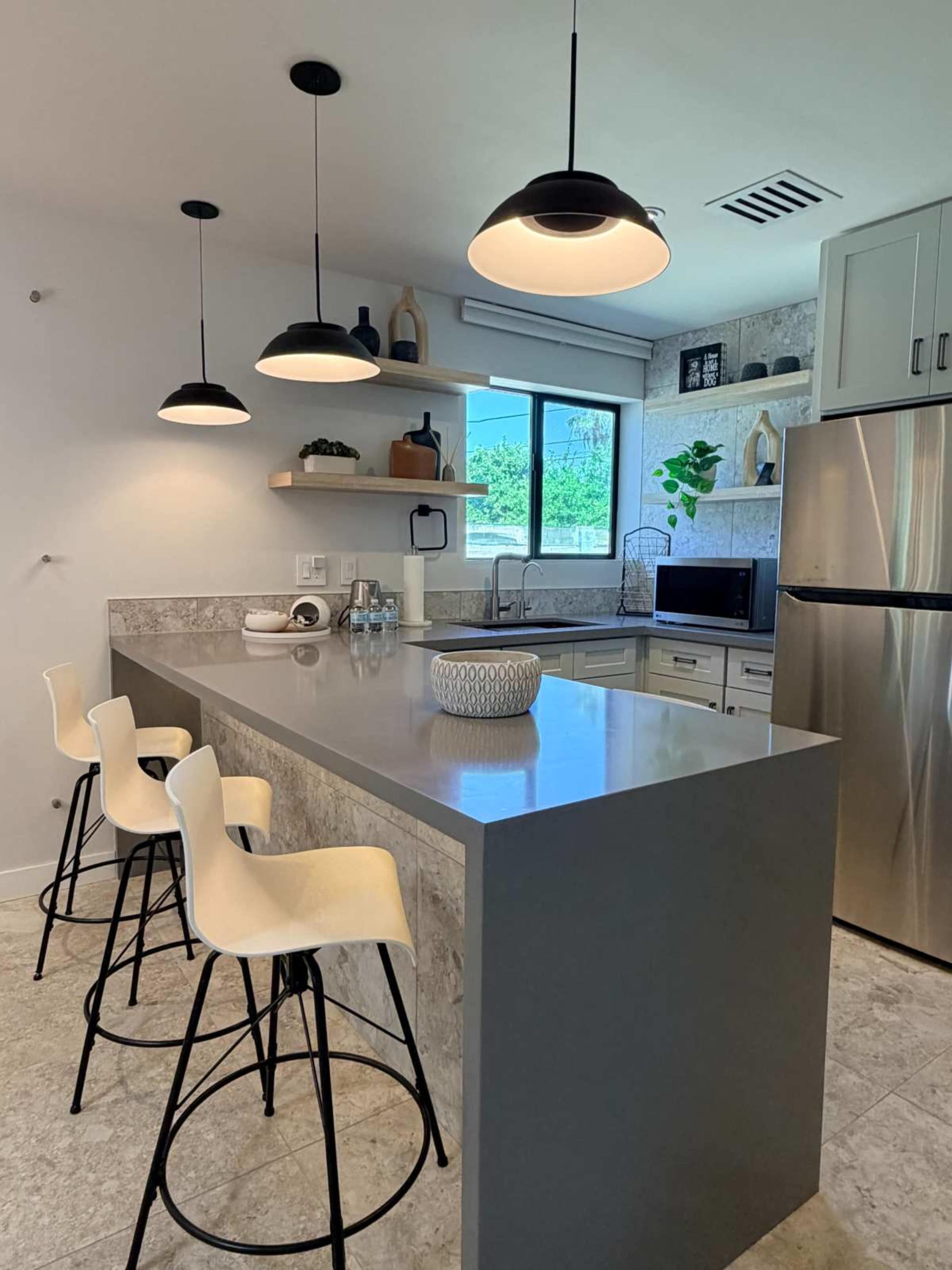 A modern kitchen features a gray island with three bar stools, overhead pendant lighting, and open shelves displaying various items.