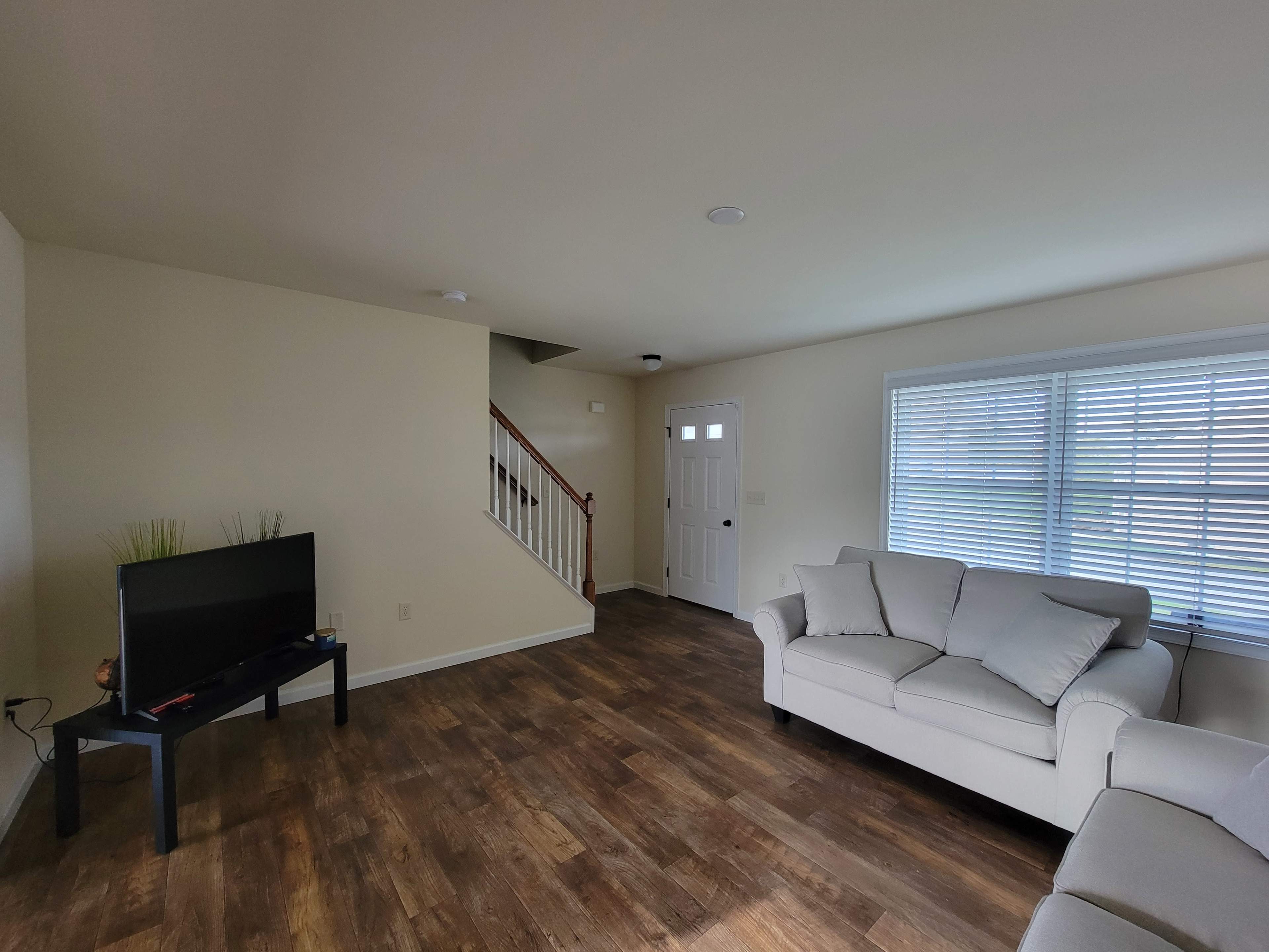 A living room featuring a light-colored sofa, a television on a stand, wooden flooring, and a staircase leading to an upper level.