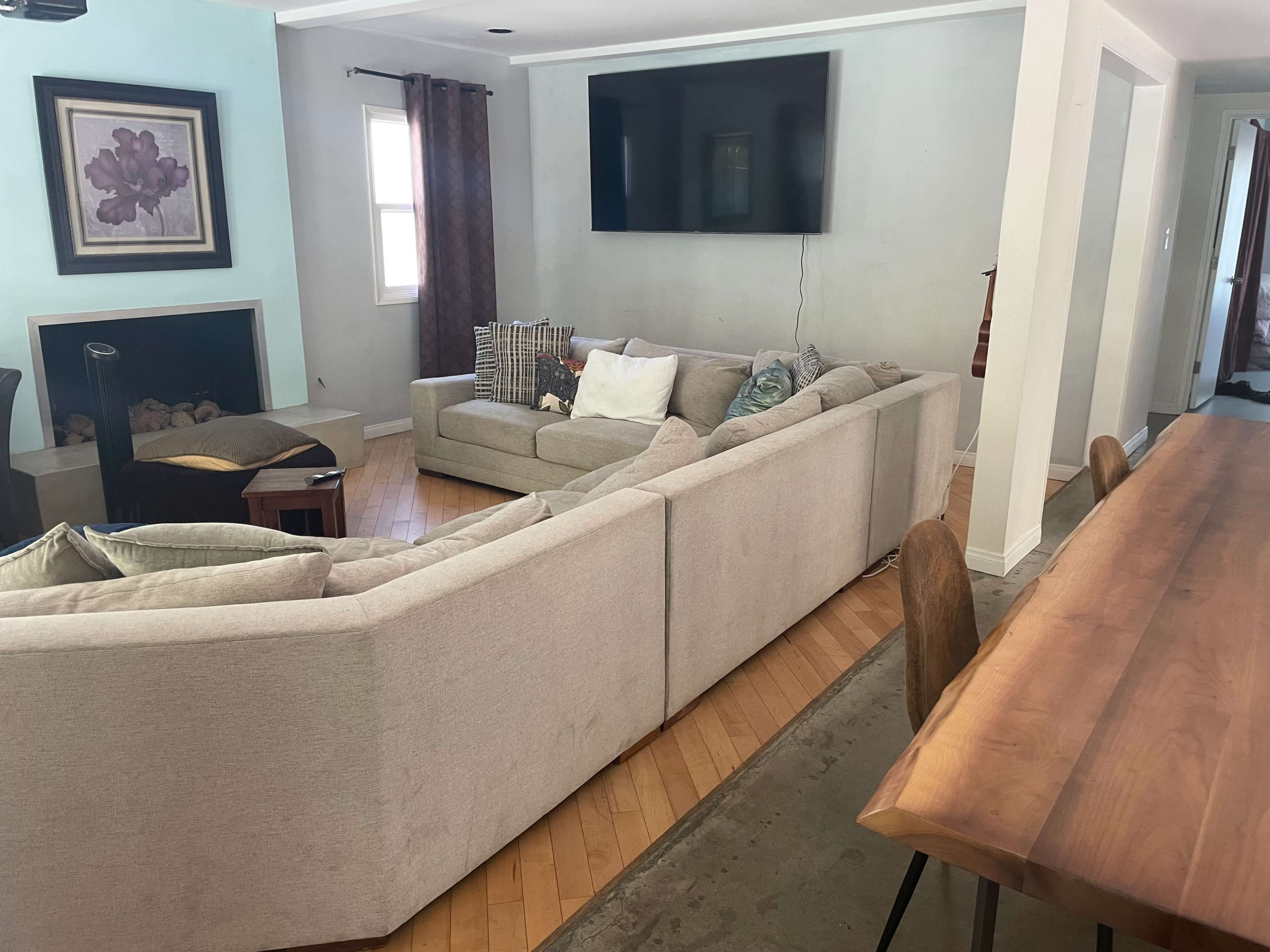 The image shows a living room with a sectional sofa, a coffee table, a wall-mounted television, and a dining area featuring a wooden table and chairs.