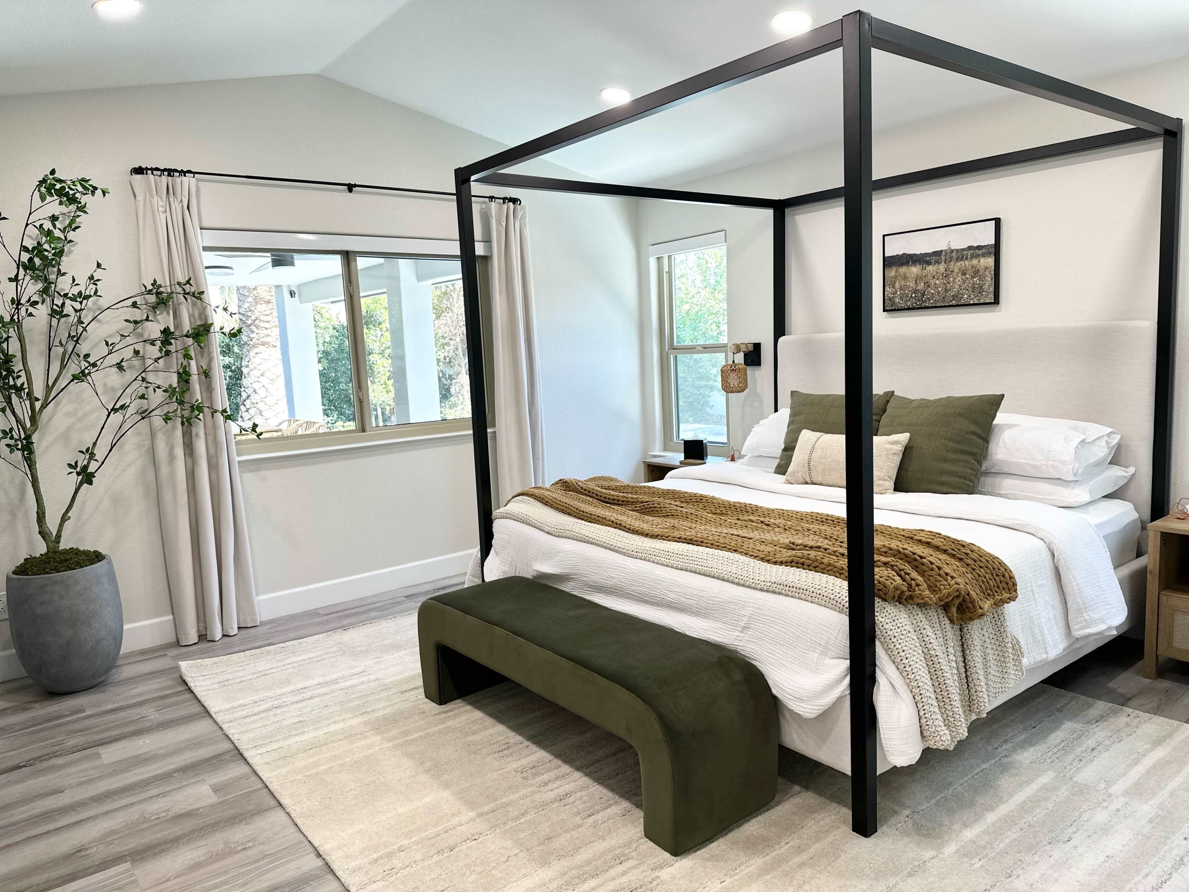 A modern bedroom featuring a four-poster bed with white bedding and a brown throw blanket, a green bench at the foot of the bed, and a large window with sheer curtains.