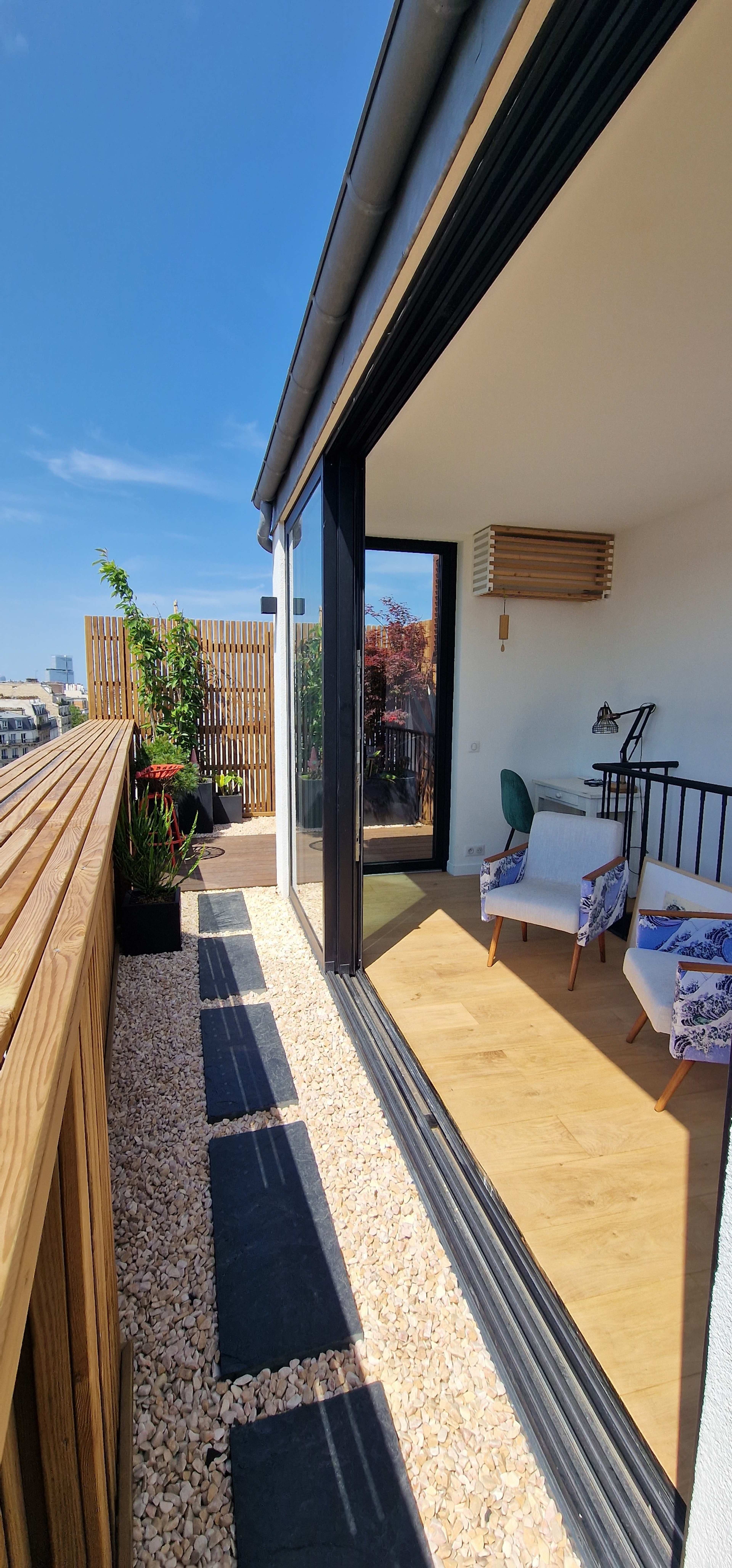 A modern balcony with a wooden railing and paved walkway leads to an indoor space featuring a chair and desk, with clear skies in the background.