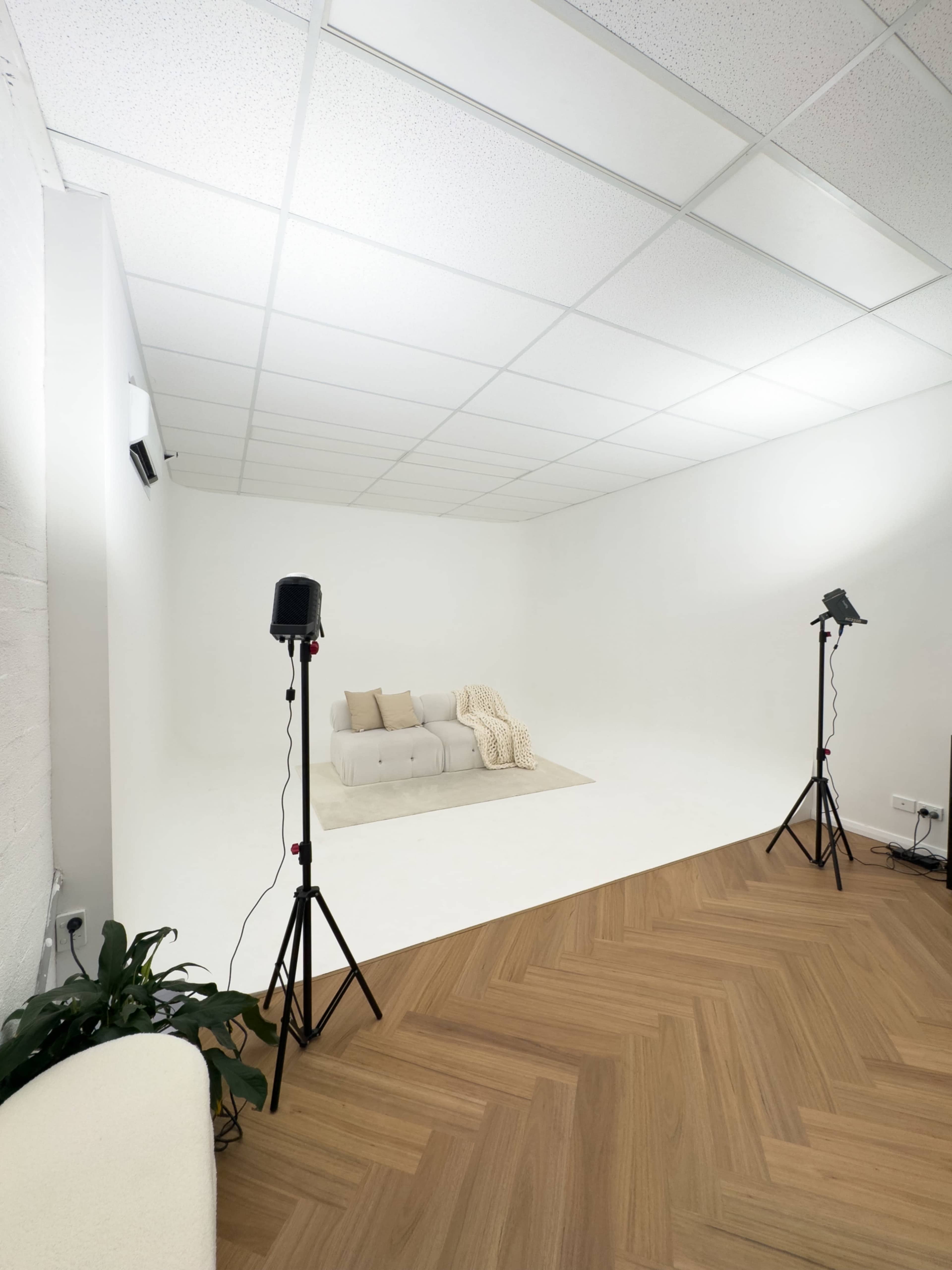 A clean, minimalistic studio space features a white backdrop, a light-colored sofa with cushions and a throw, and two standing lights positioned nearby.