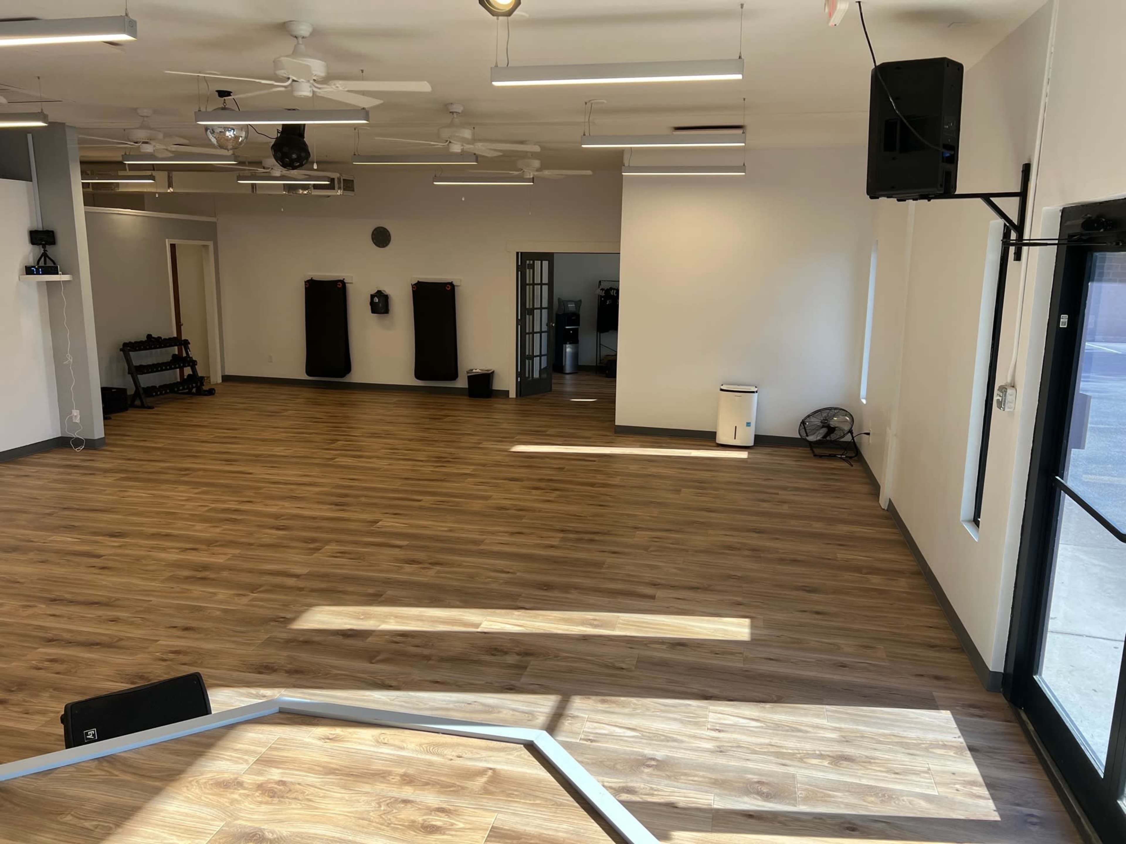 A spacious, empty room with wooden flooring, ceiling fans, and several fitness equipment stations along the walls.