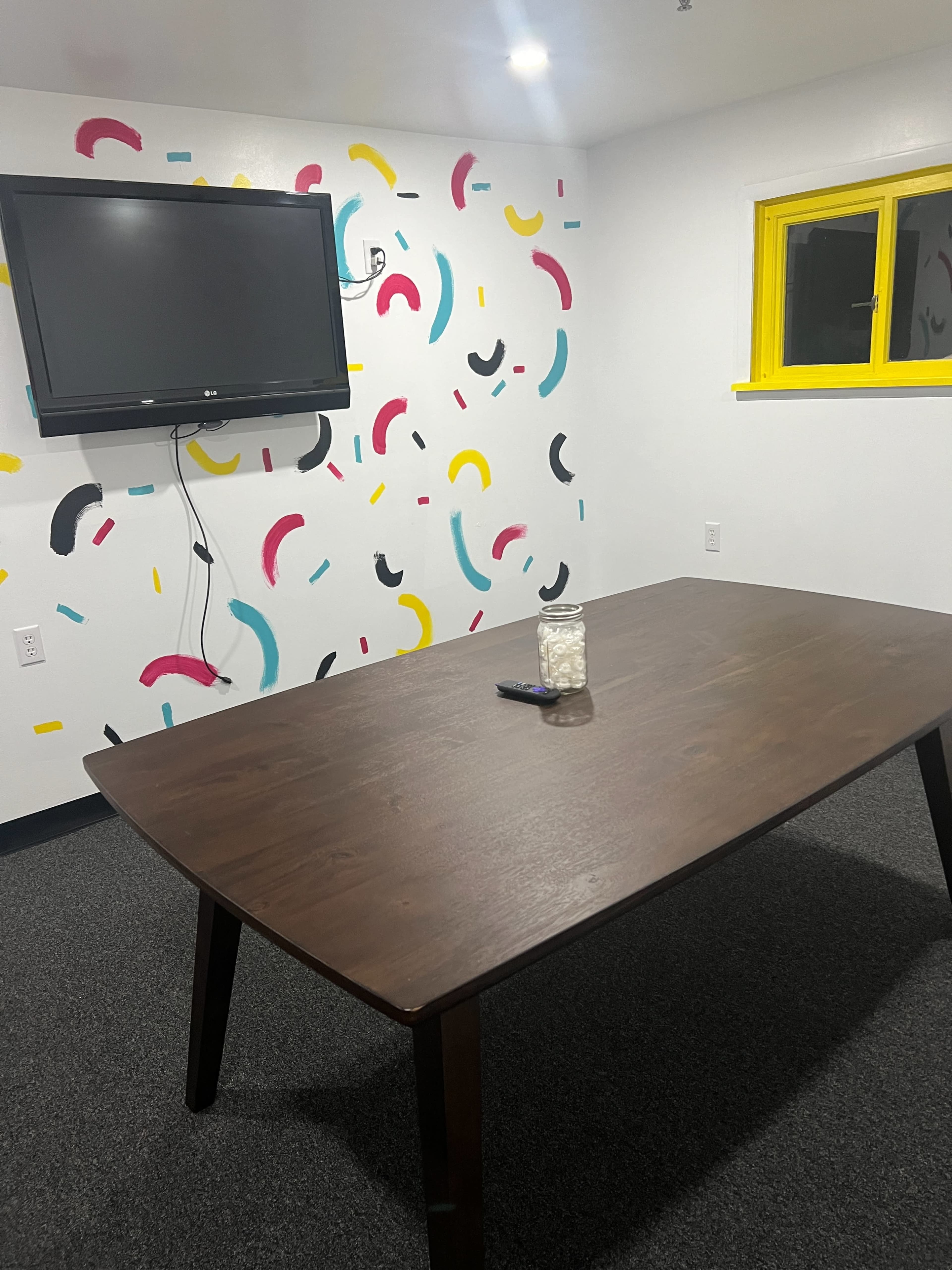 A conference room features a wooden table, a television mounted on the wall, and a colorful abstract mural behind it.