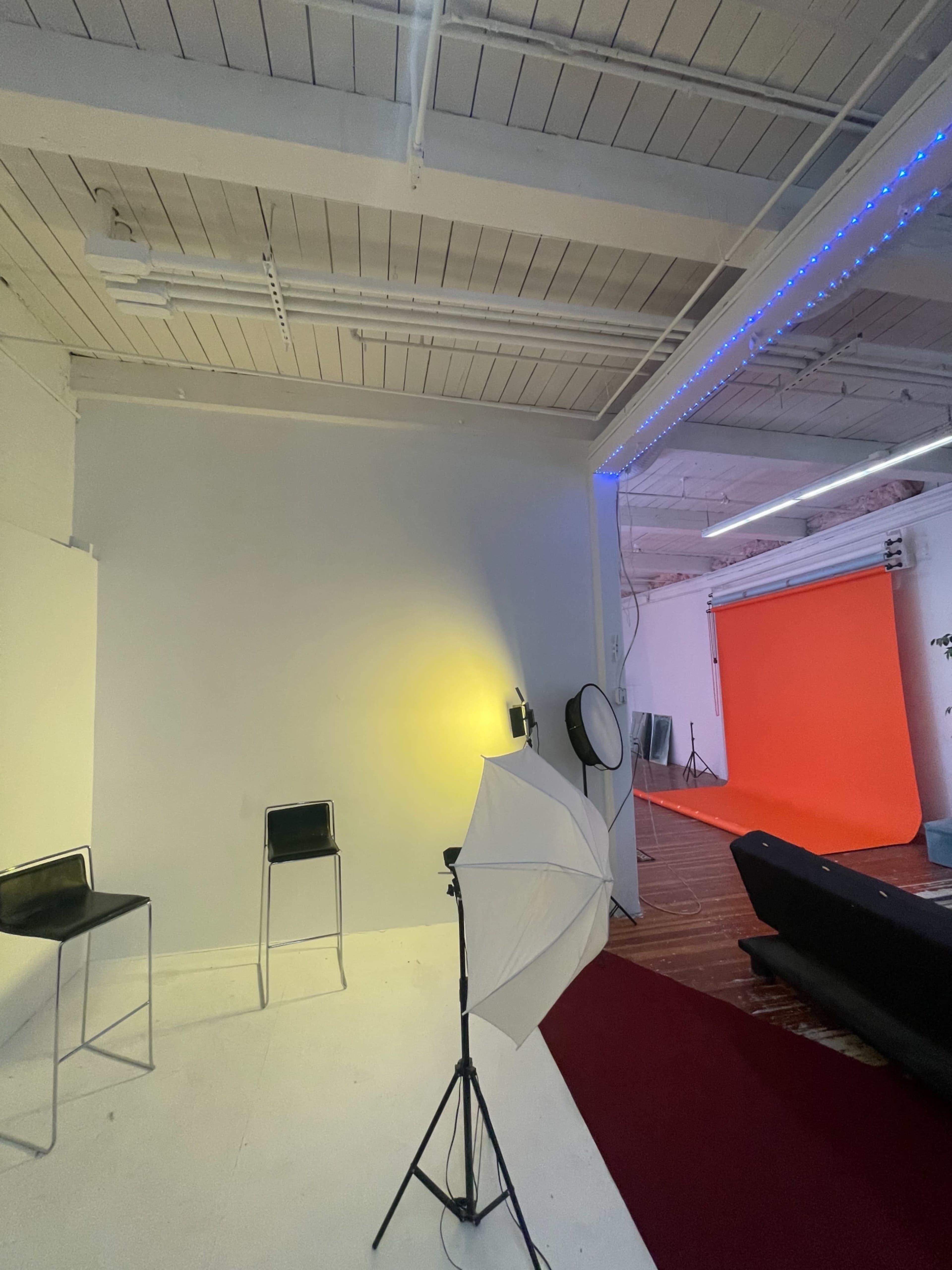 The image shows a photography studio with a simple white backdrop, two black chairs, a lighting setup, and an orange backdrop in a separate area.