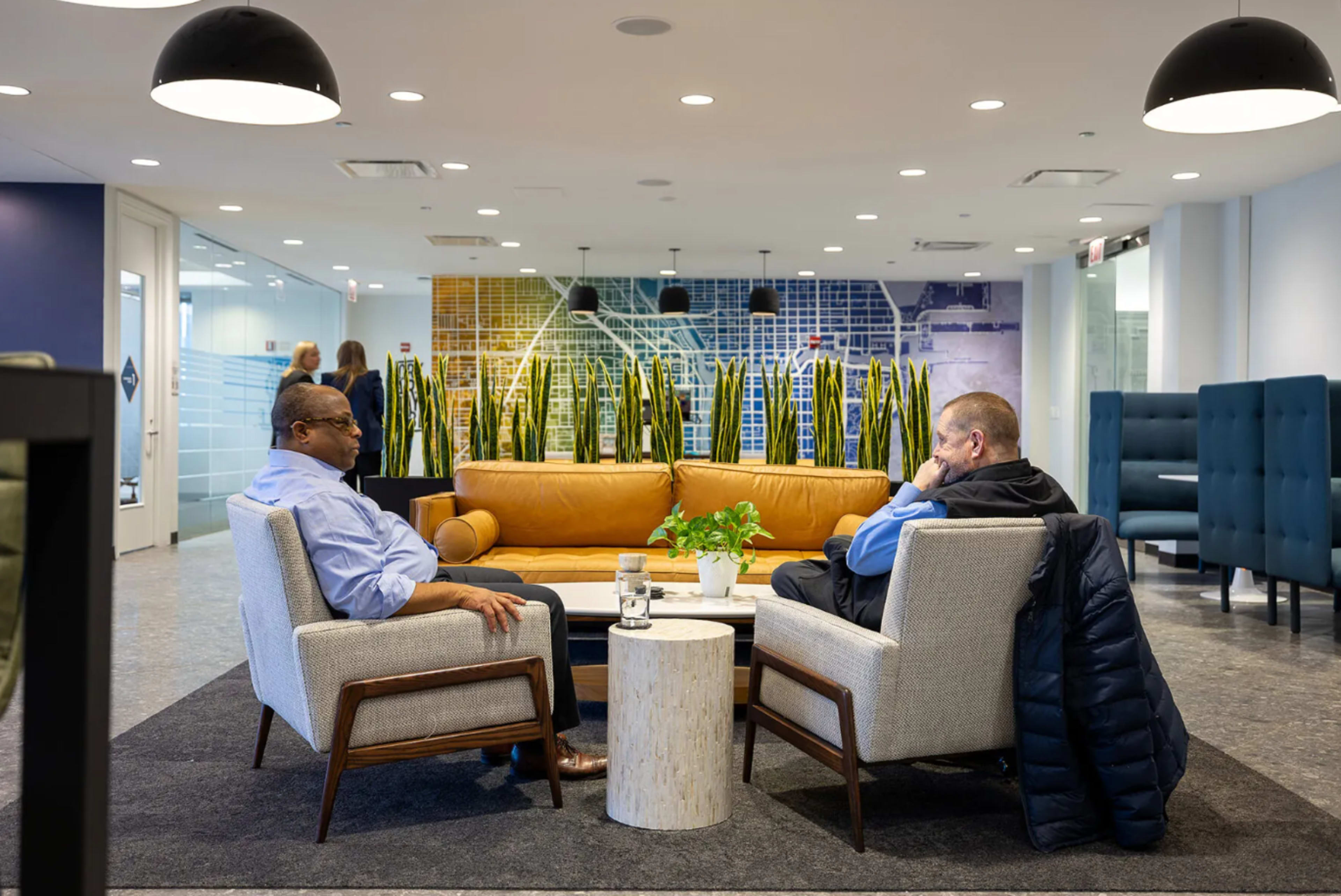 Two men sit across from each other in a modern office lounge, surrounded by decorative plants and contemporary furniture.