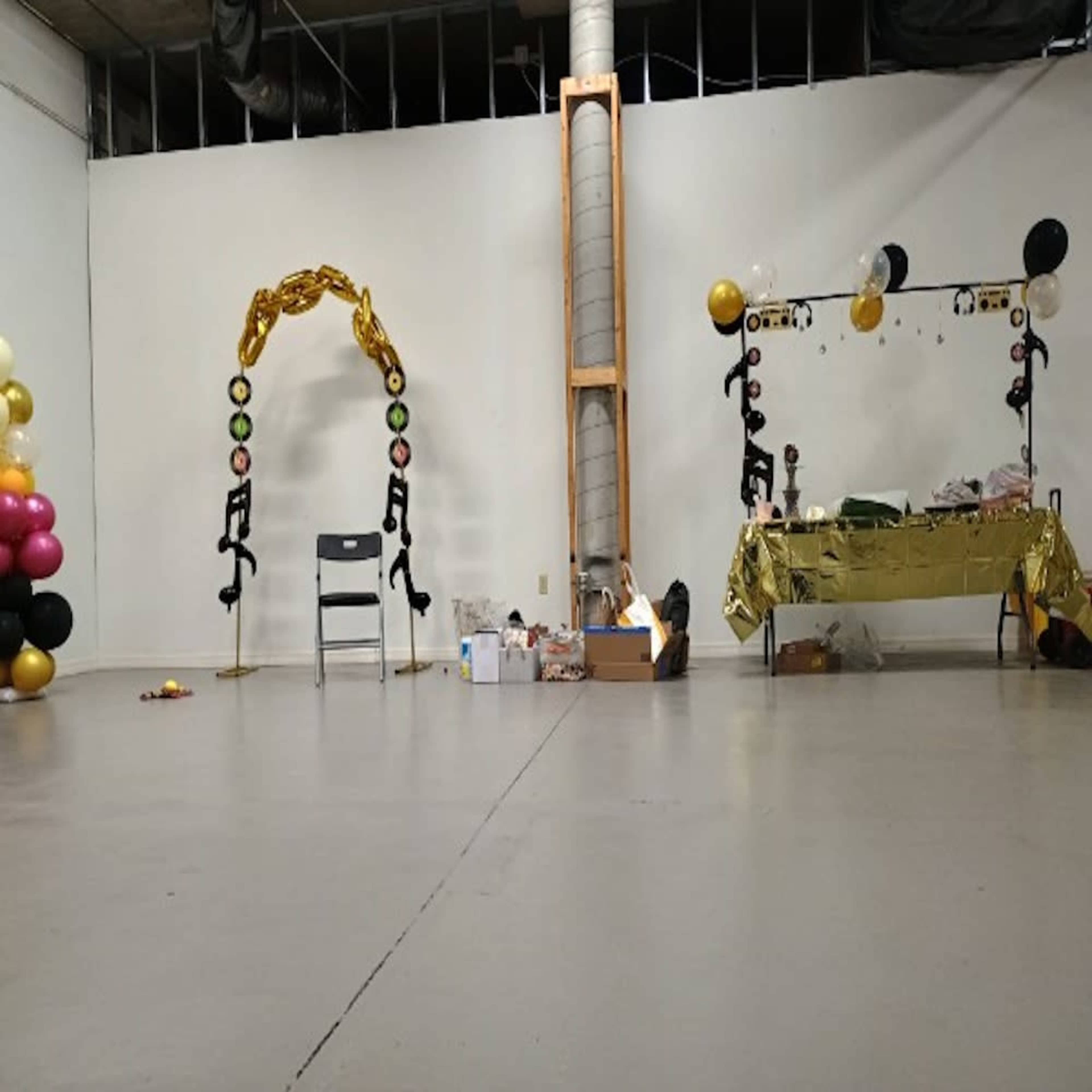 The image shows an empty indoor space with a gold-themed decoration setup on one side, including a table and an arch adorned with balloons and other decor.