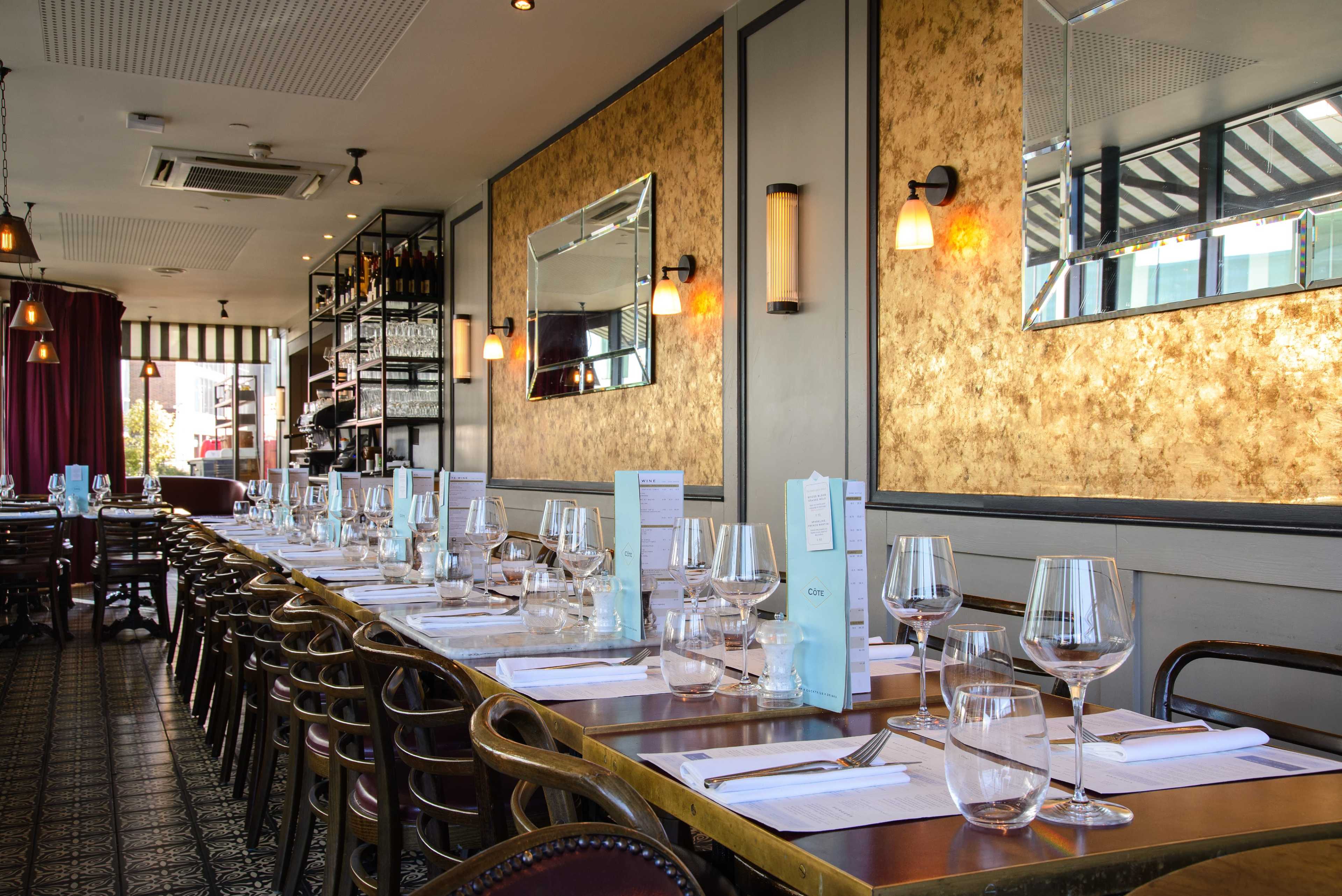 The image shows a restaurant interior with neatly set tables featuring glasses and menus, along with decorative wall accents and ambient lighting.