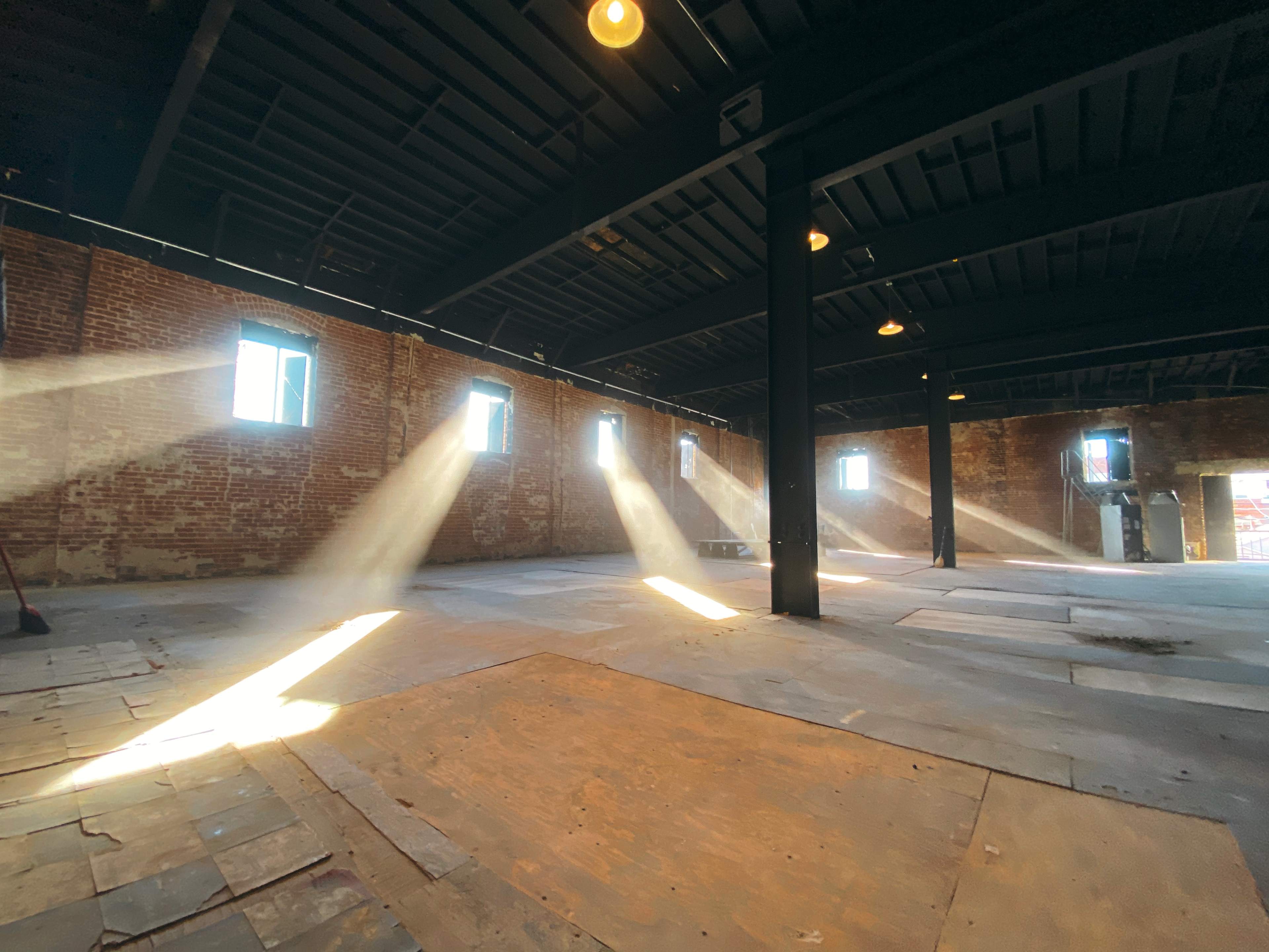 Sunlight streams through windows onto the floor of an empty brick room with a black ceiling and exposed beams.