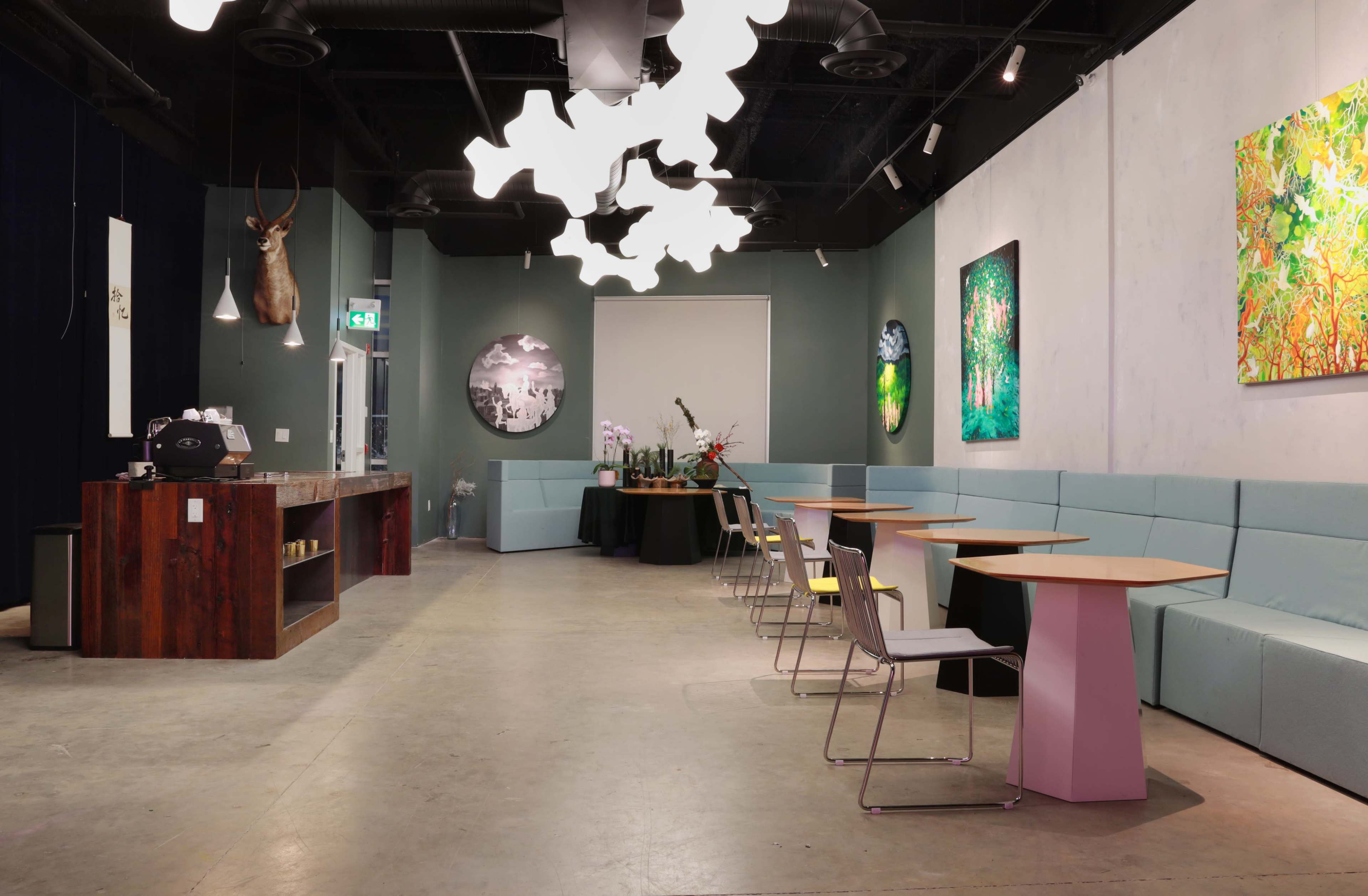 The interior of a modern cafe features abstract light fixtures, a wooden counter, and art pieces on the walls, with seating along one side and tables arranged in the middle.