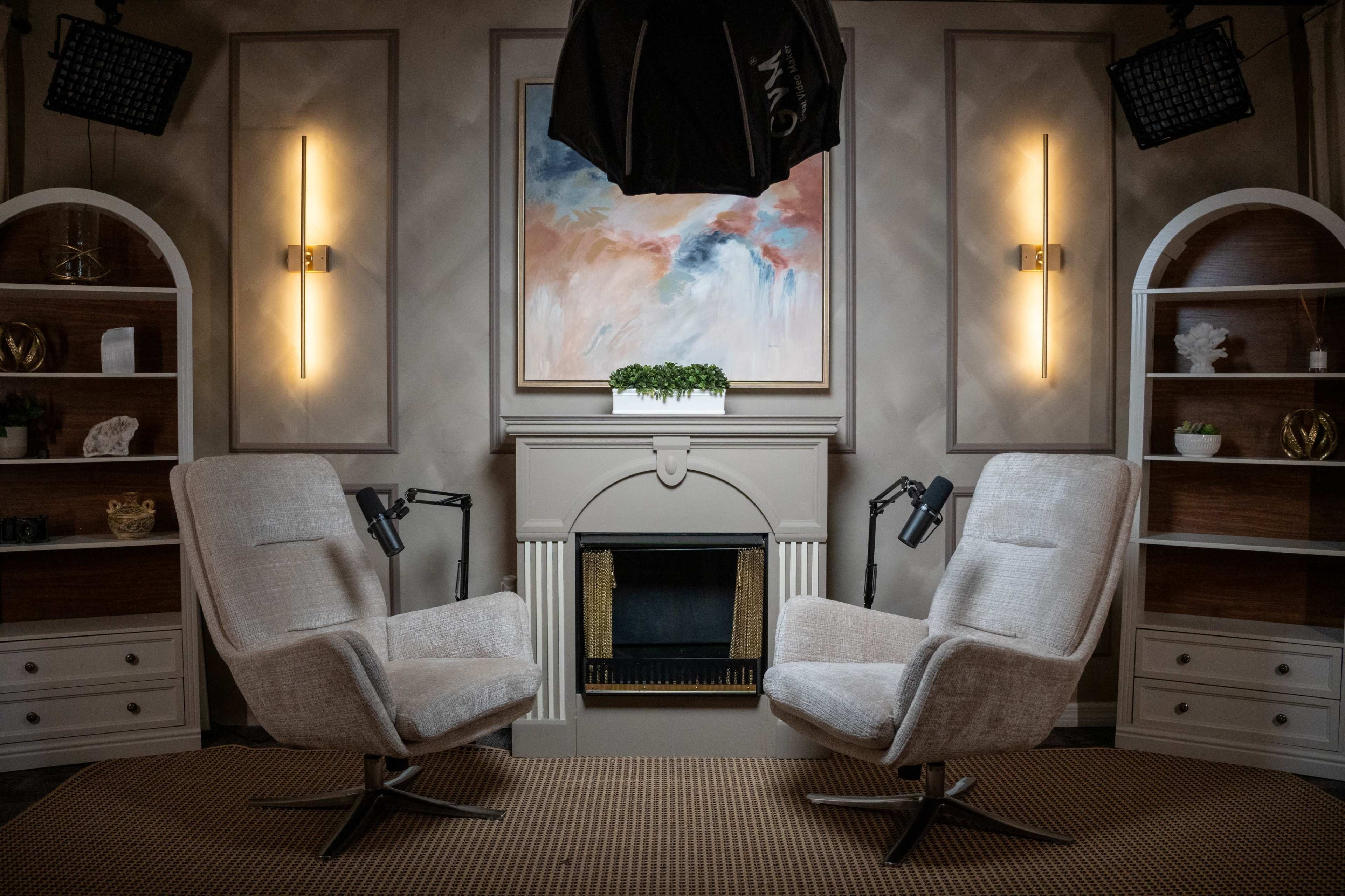 The image shows a well-decorated studio setup with two grey armchairs facing a fireplace, flanked by shelves, and illuminated by soft lighting.