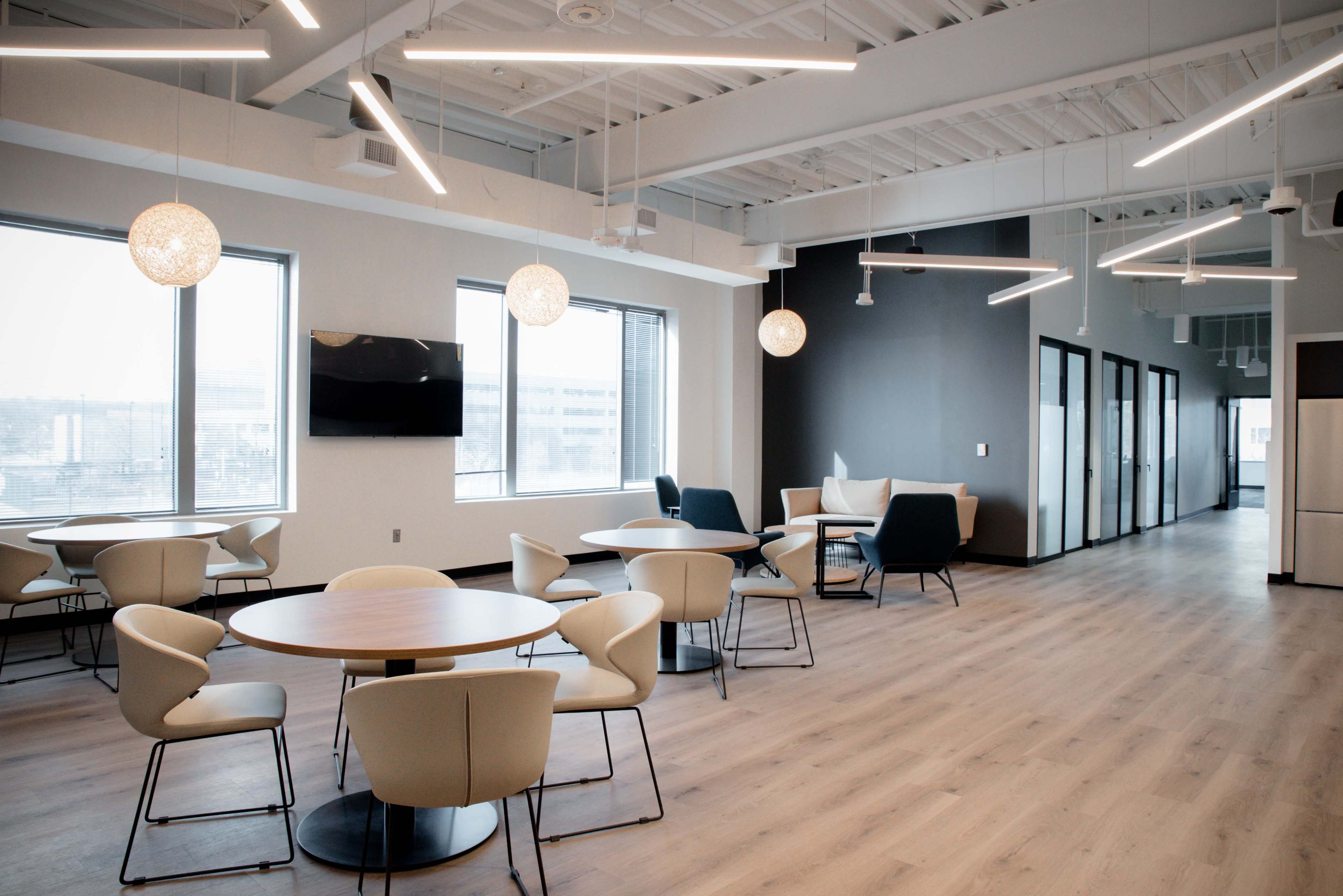 The image shows a modern office space featuring circular tables and chairs, pendant lighting, large windows, and glass-walled meeting rooms.