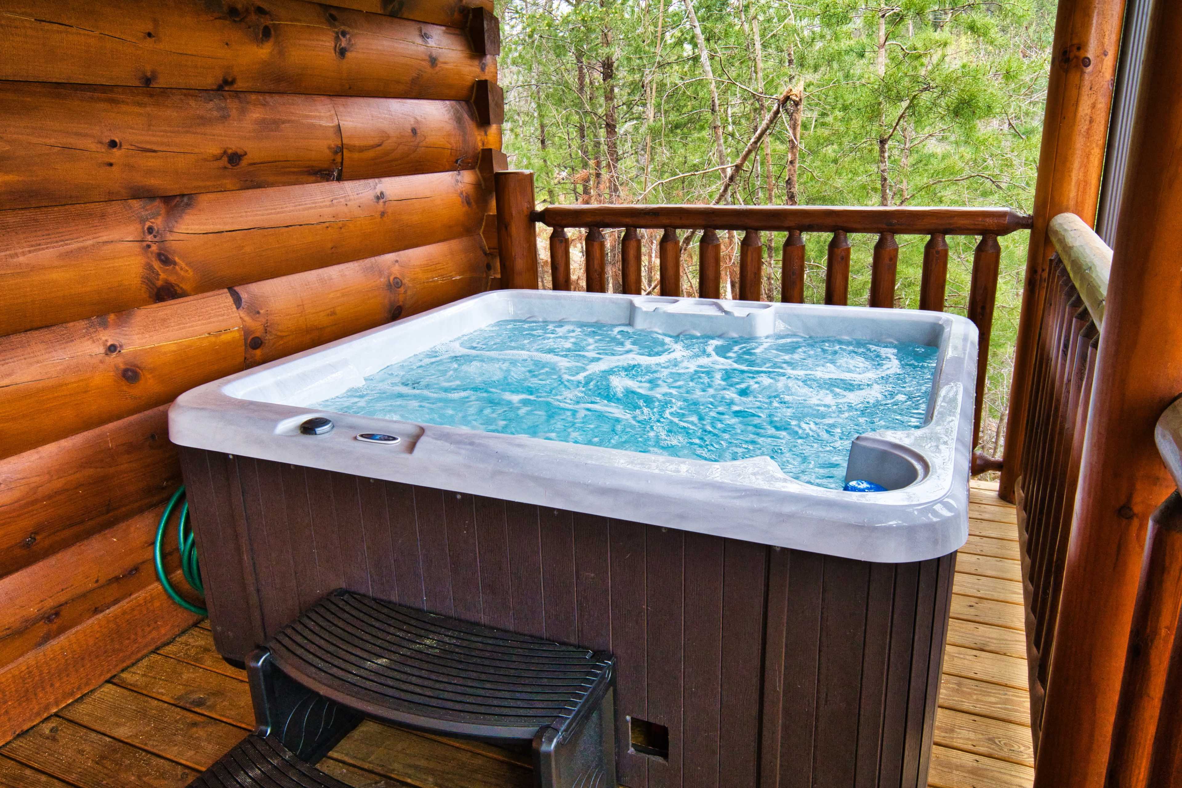 A hot tub with bubbling water is situated on a wooden deck adjacent to a log cabin, surrounded by trees.