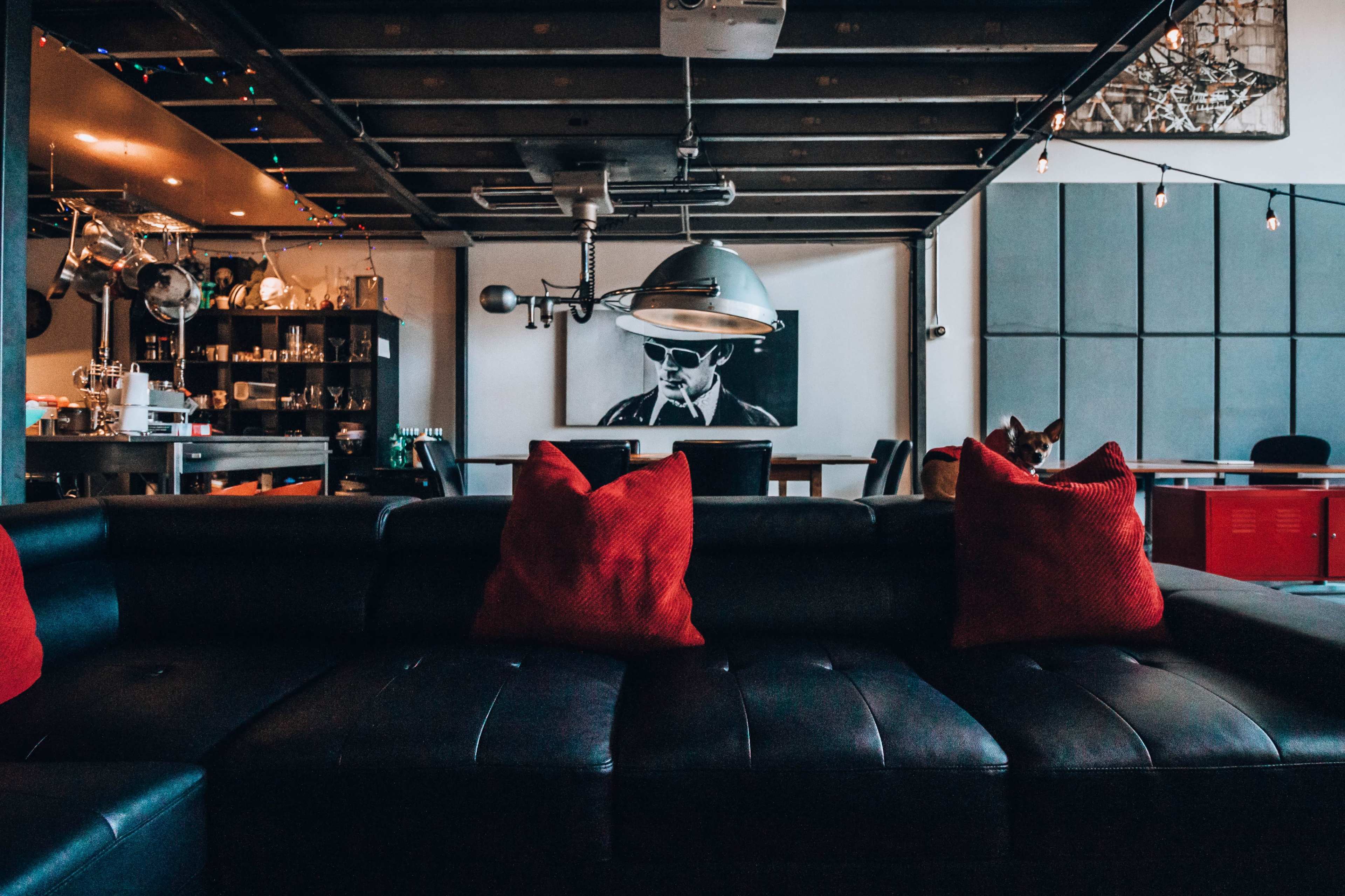 A black leather sectional couch with red pillows faces a wall adorned with a large black-and-white portrait and a modern dining area in the background.