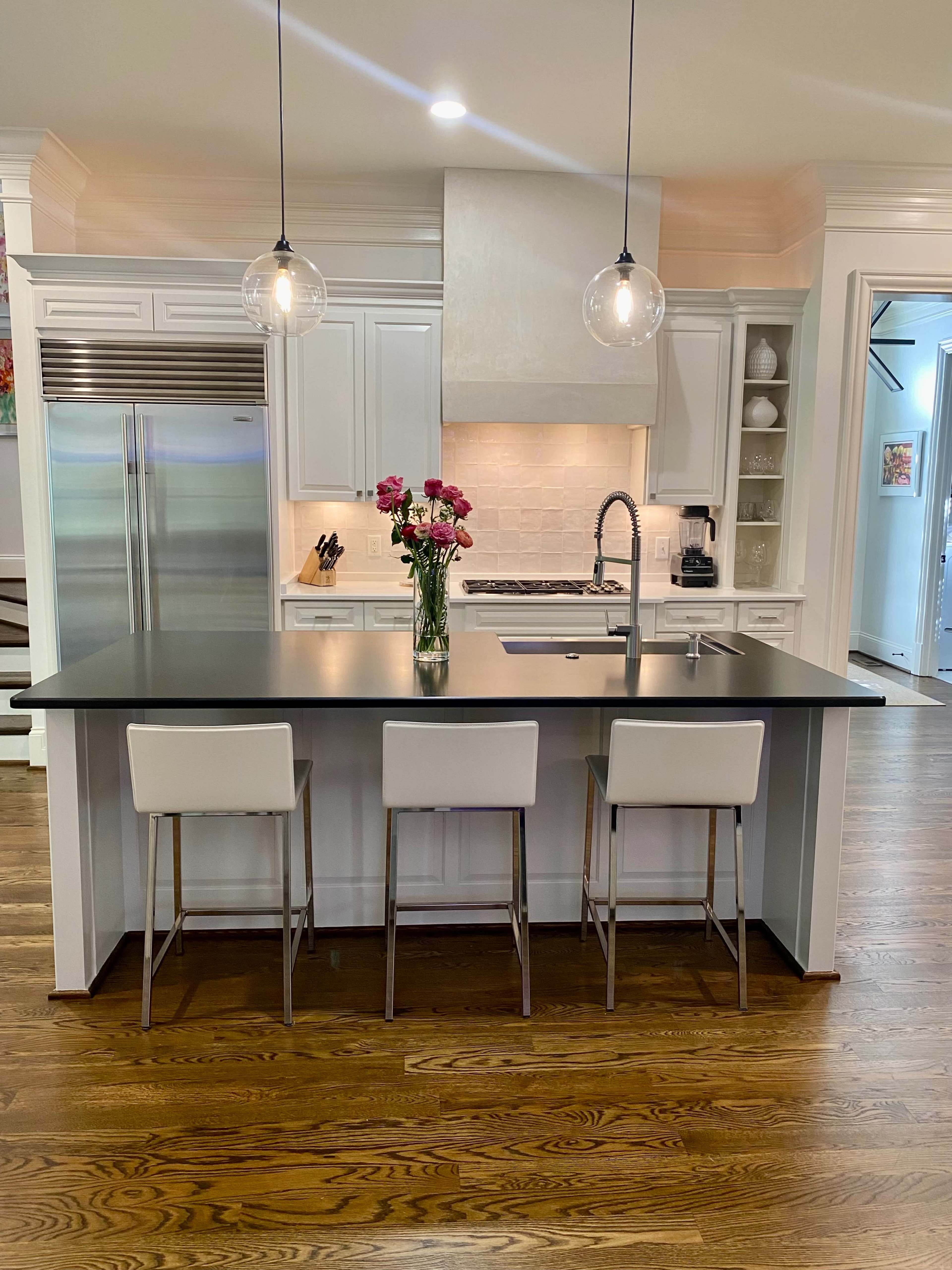 A modern kitchen features a central island with three stools, a stainless steel refrigerator, and pendant lights above a sink.