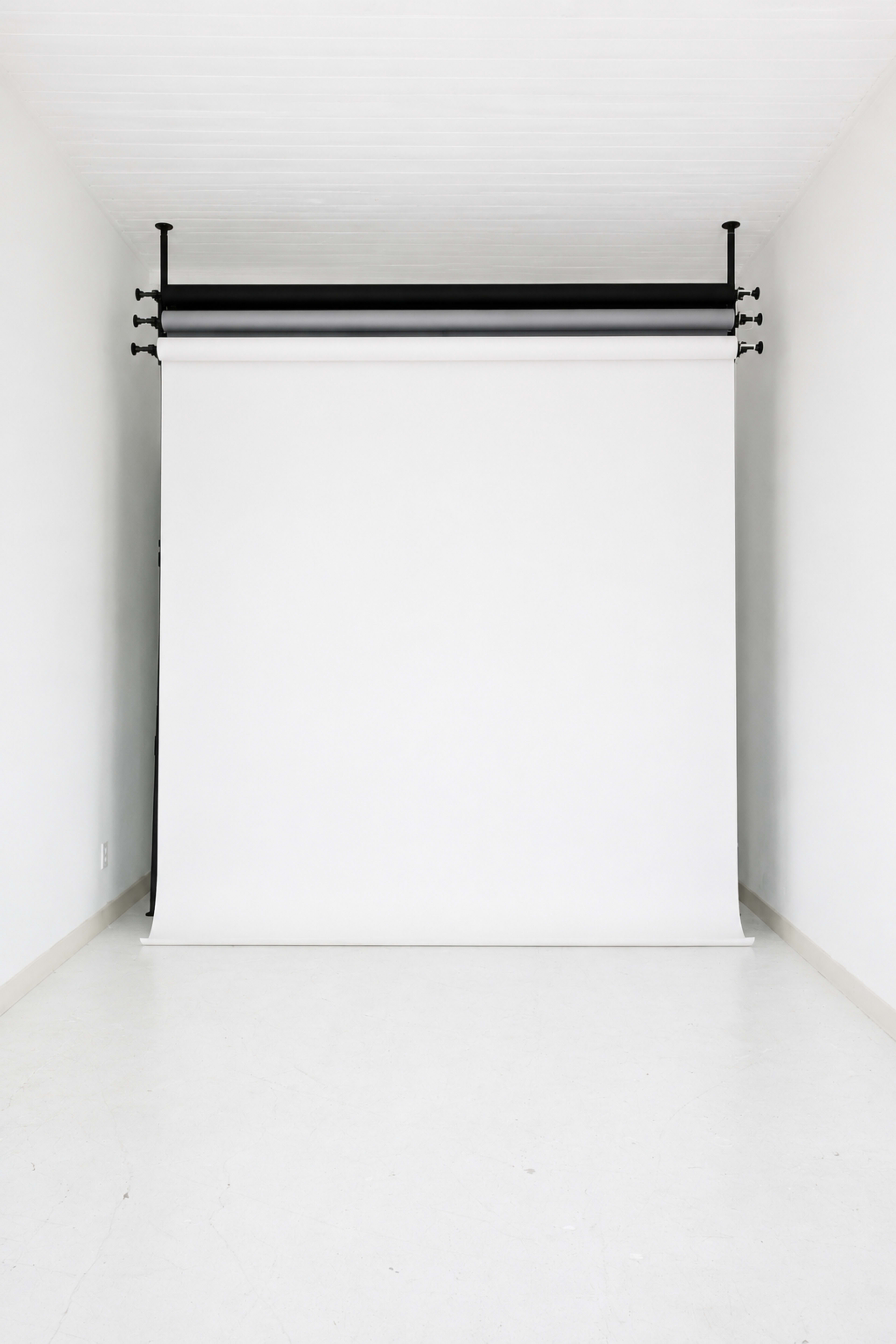 The image shows an empty studio space with a white backdrop hanging from a black metal support structure against a plain wall.