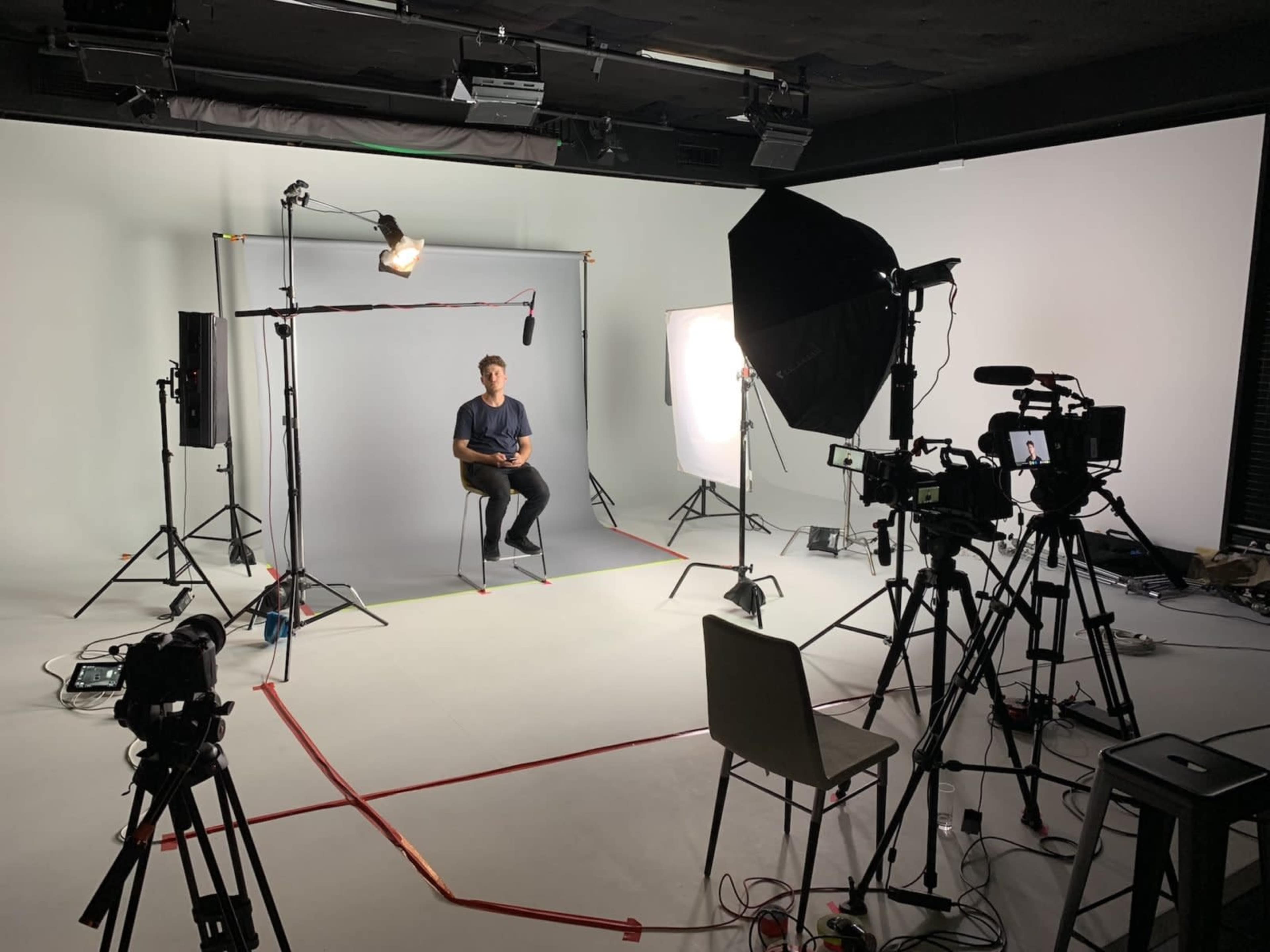 A man sits on a stool in a studio set up for filming, surrounded by multiple cameras and lighting equipment.