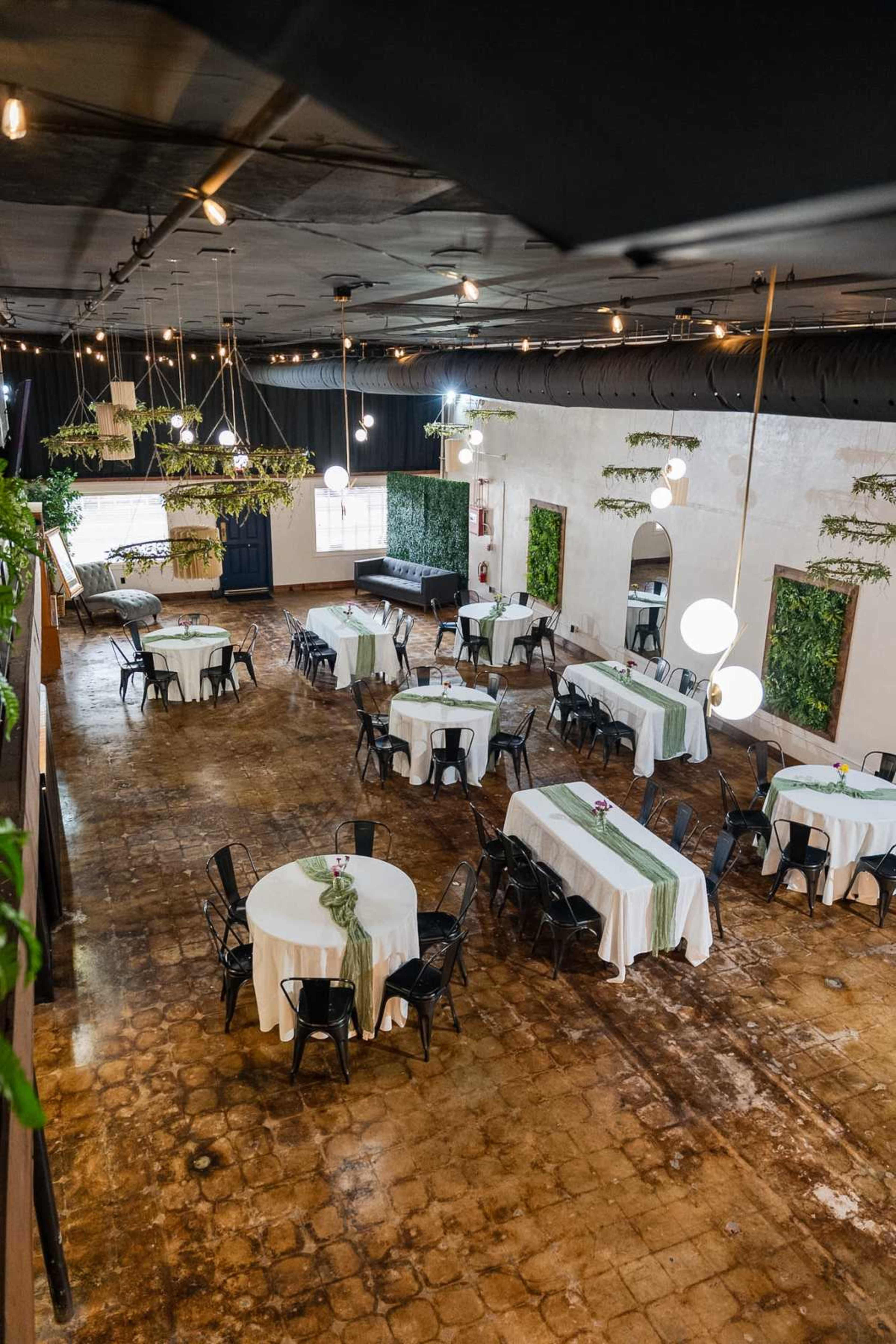The image shows a spacious indoor event venue with round tables set for an occasion, featuring white tablecloths and black chairs, and greenery decor along the walls.