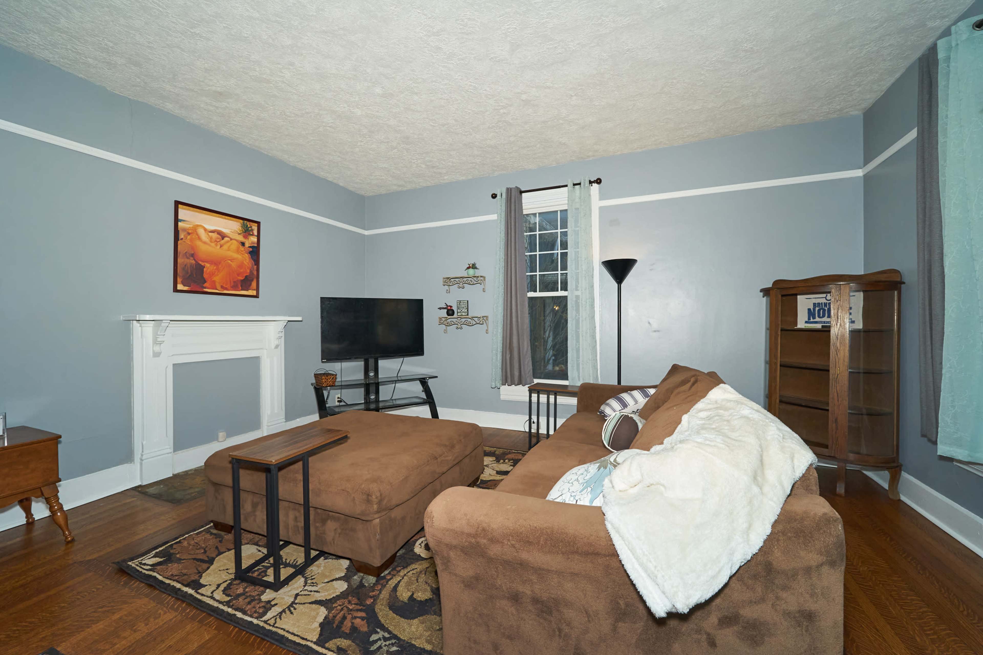 The living room features a brown sectional sofa, a television on a stand, a wooden bookshelf, and a decorative painting on the wall.