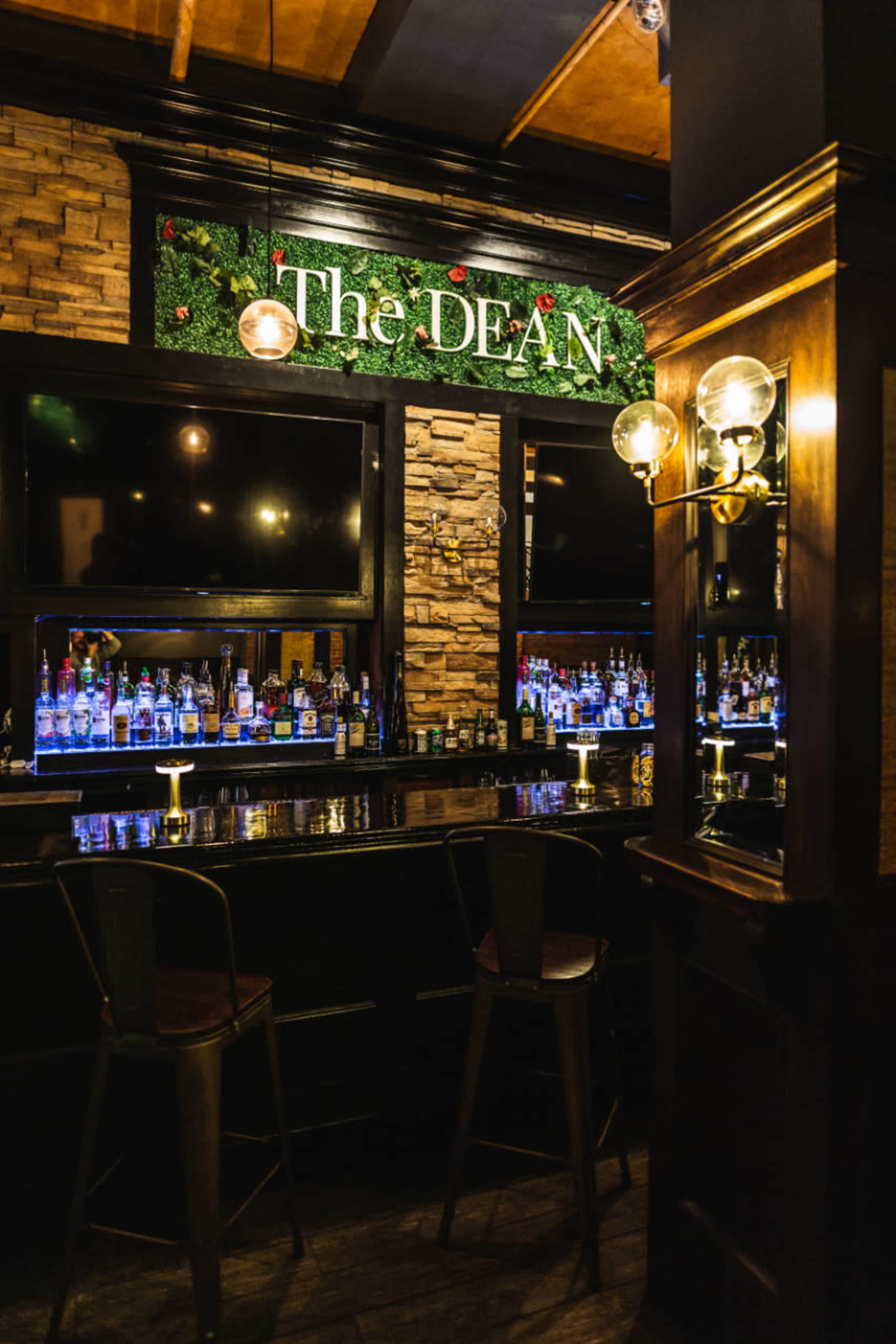 The image shows a dimly lit bar area with a wooden counter, shelves lined with various bottles, and a sign reading "The DEAN" decorated with greenery.