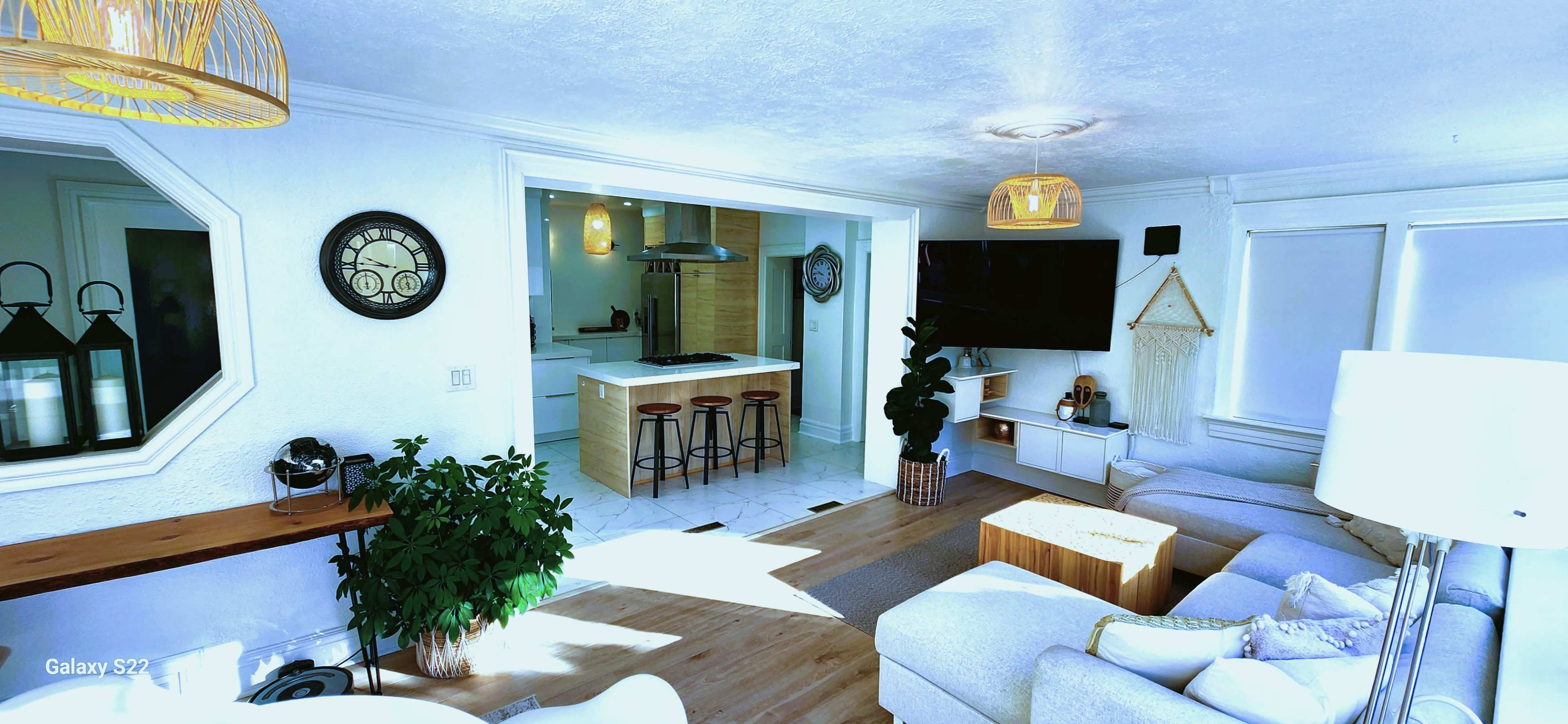 A bright living room that connects to a kitchen, featuring modern furniture, a large television, and plants.