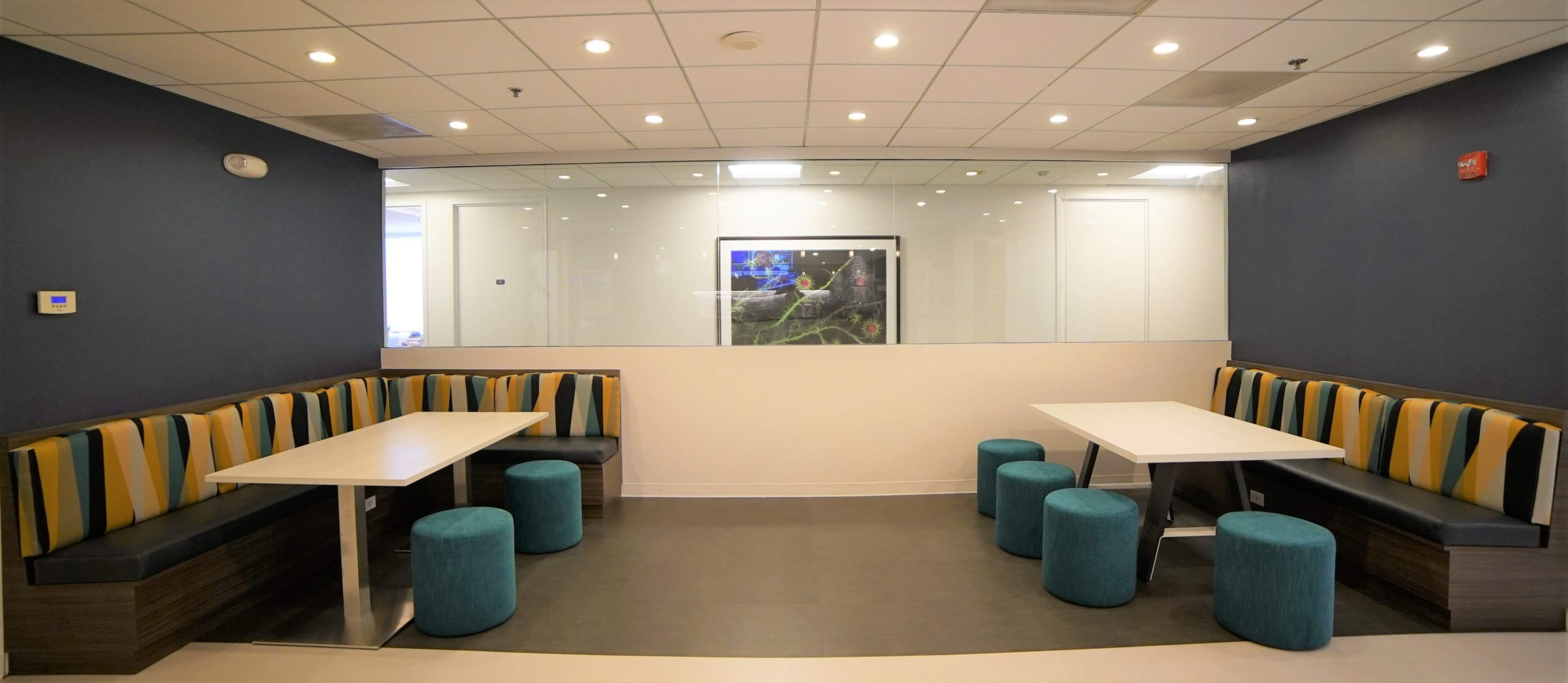 A modern seating area with two tables and bench seating, surrounded by blue and yellow striped cushions, along with teal stools in front of a glass wall.