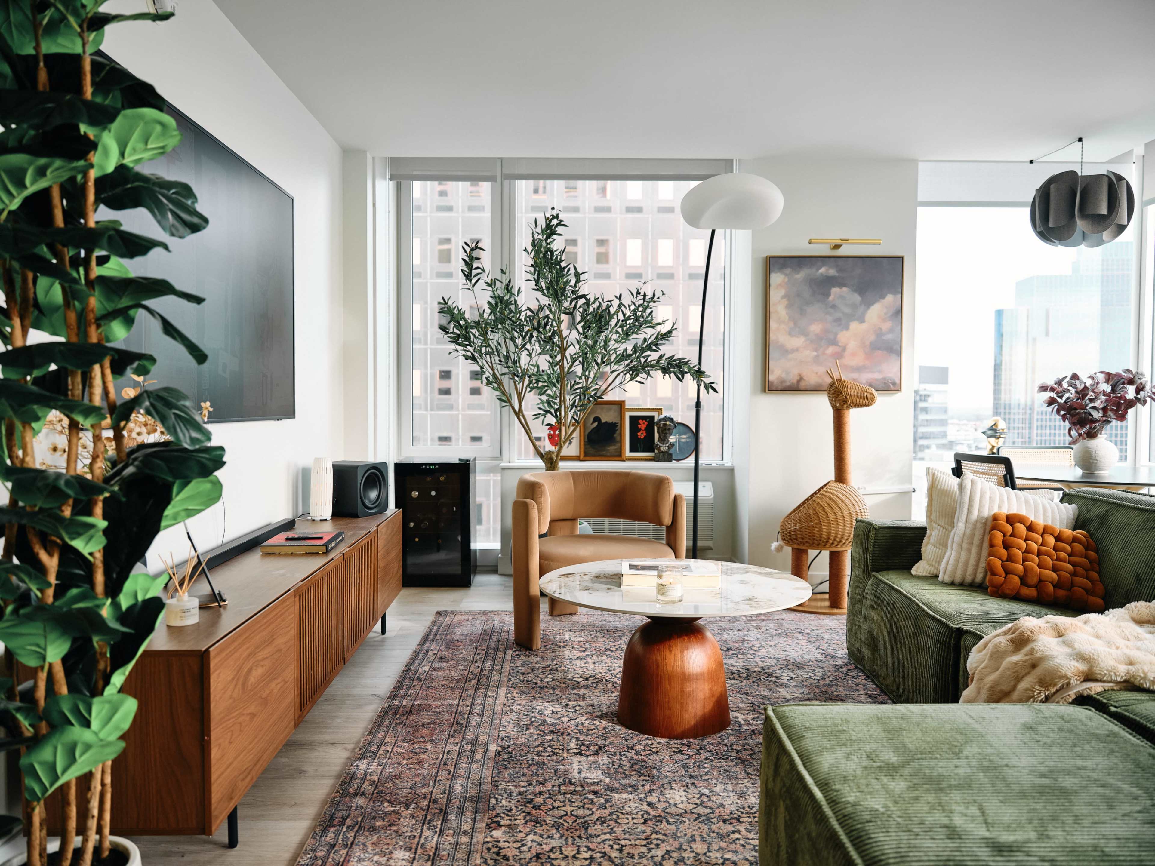 The image shows a modern living room with green upholstered furniture, a large wall-mounted television, and a coffee table surrounded by decorative plants and artwork.