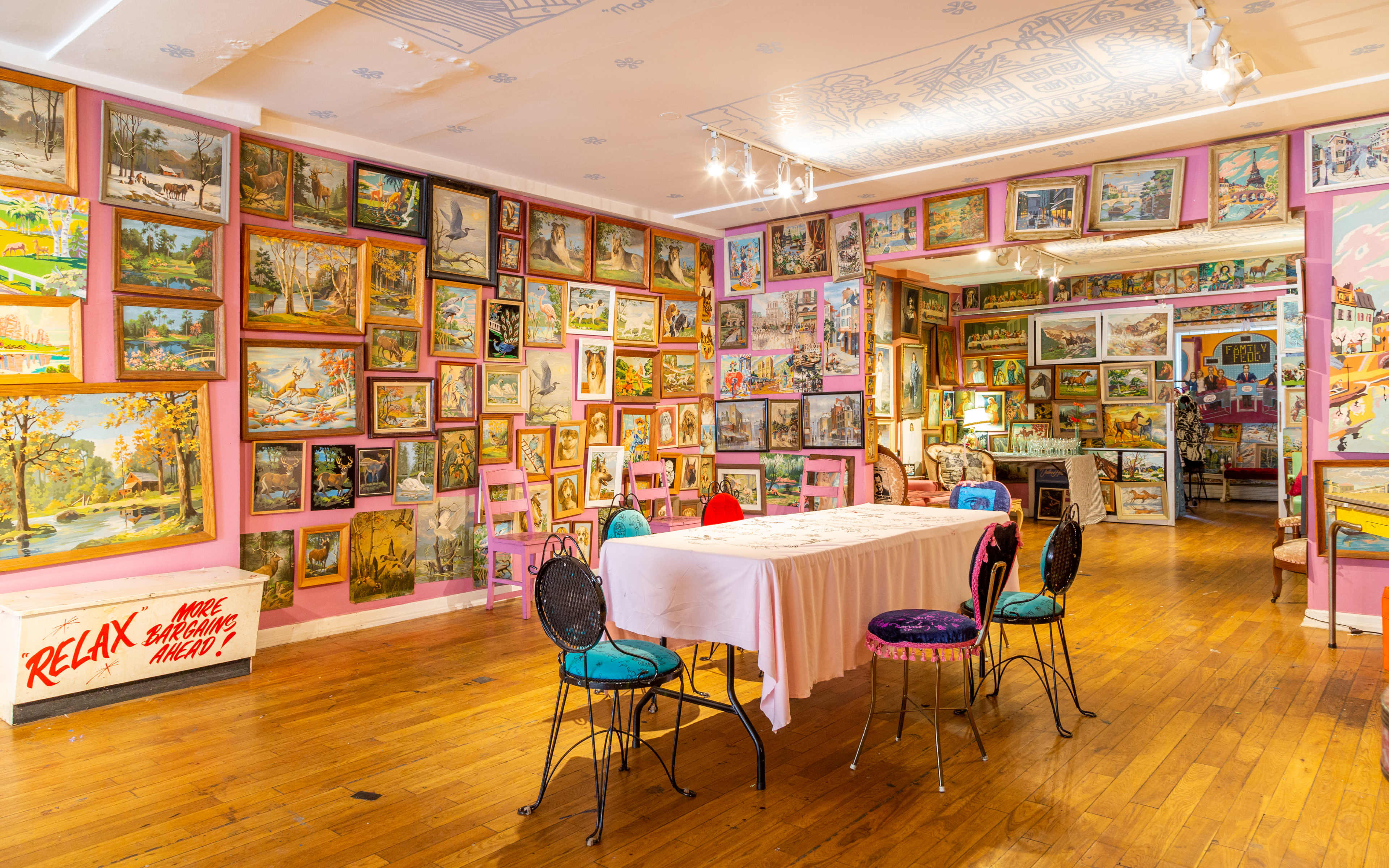 The room is filled with a variety of colorful paintings displayed on the walls, with a central table surrounded by chairs.