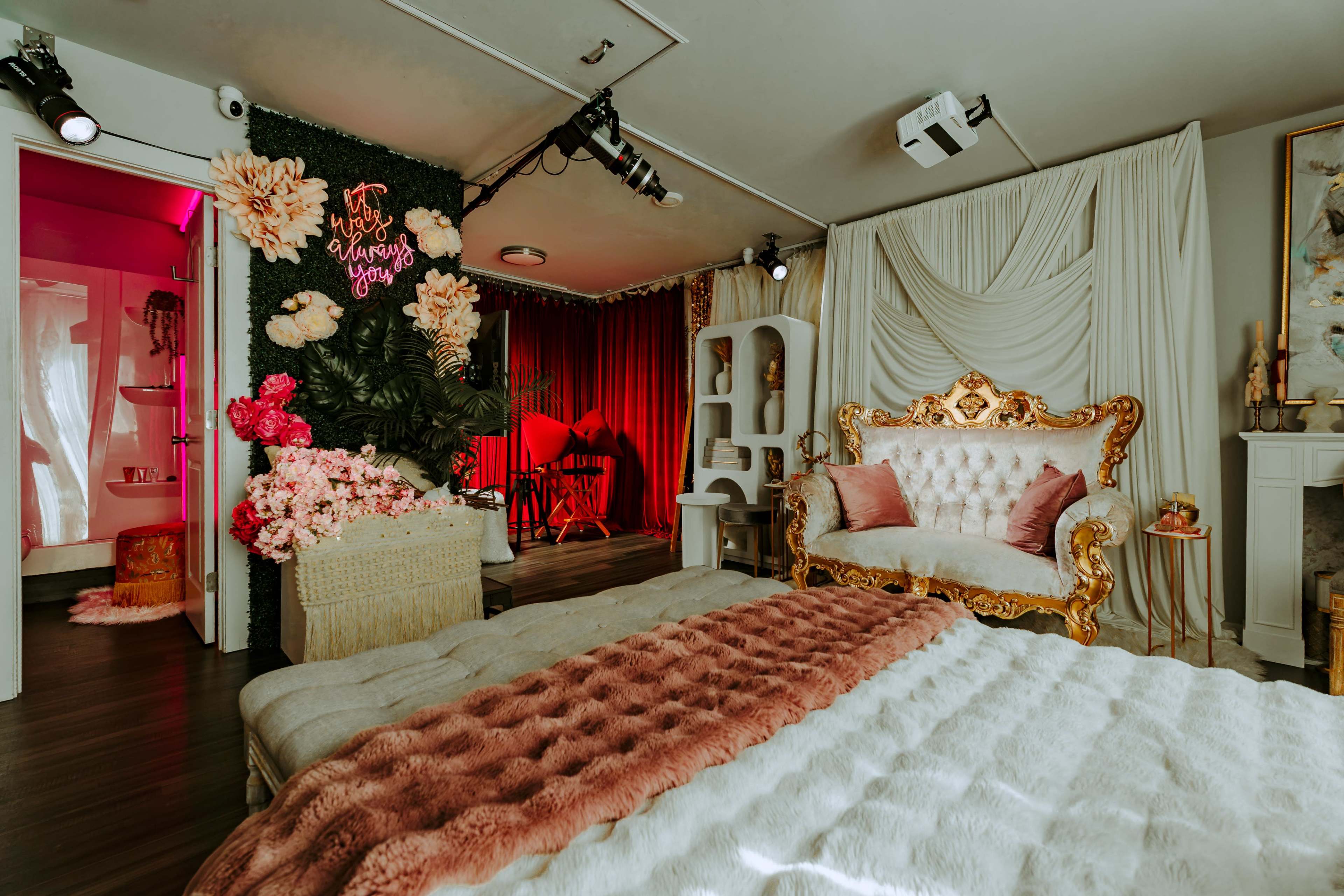 The image depicts a styled room featuring a plush bed, a decorative sofa, and floral arrangements against a backdrop of curtains and lighting.