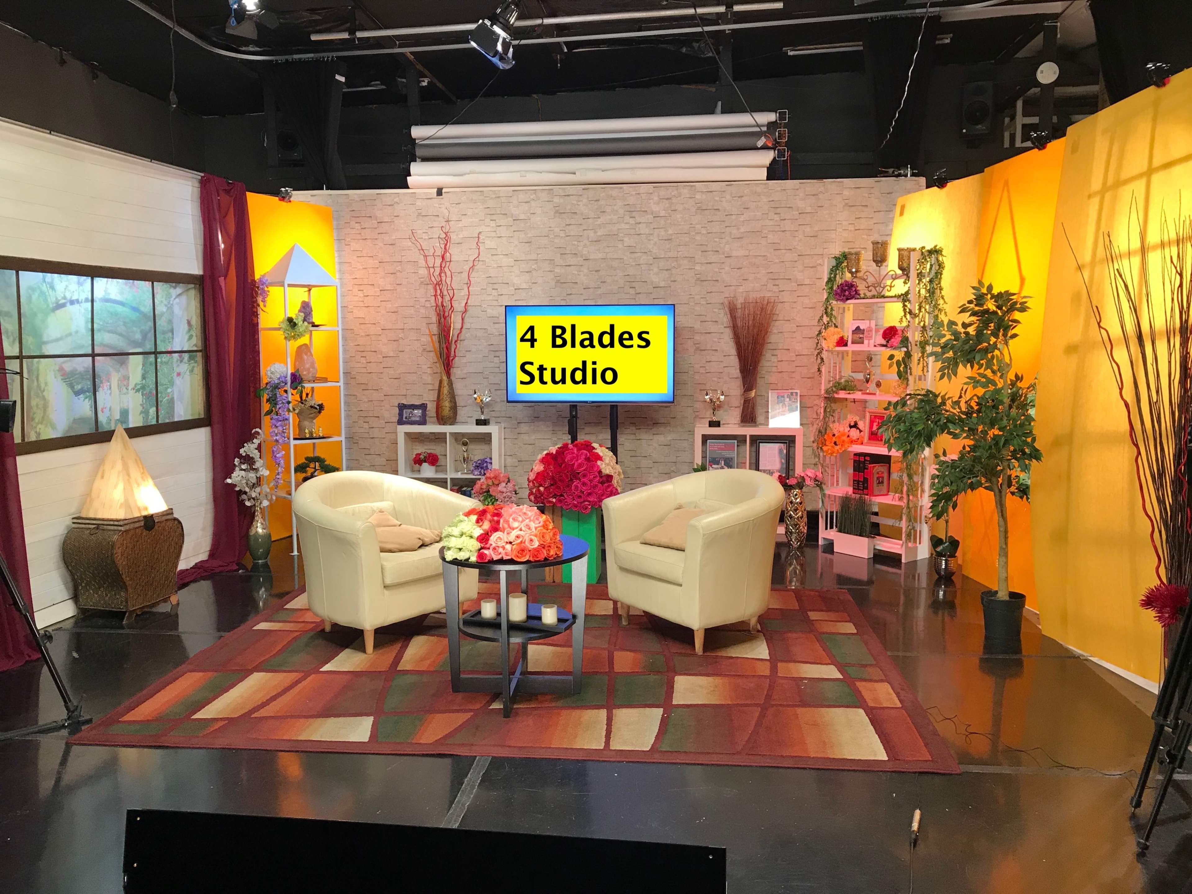 The image shows a brightly lit television studio set with two white chairs, a colorful rug, decorative plants, and a large screen labeled "4 Blades Studio."
