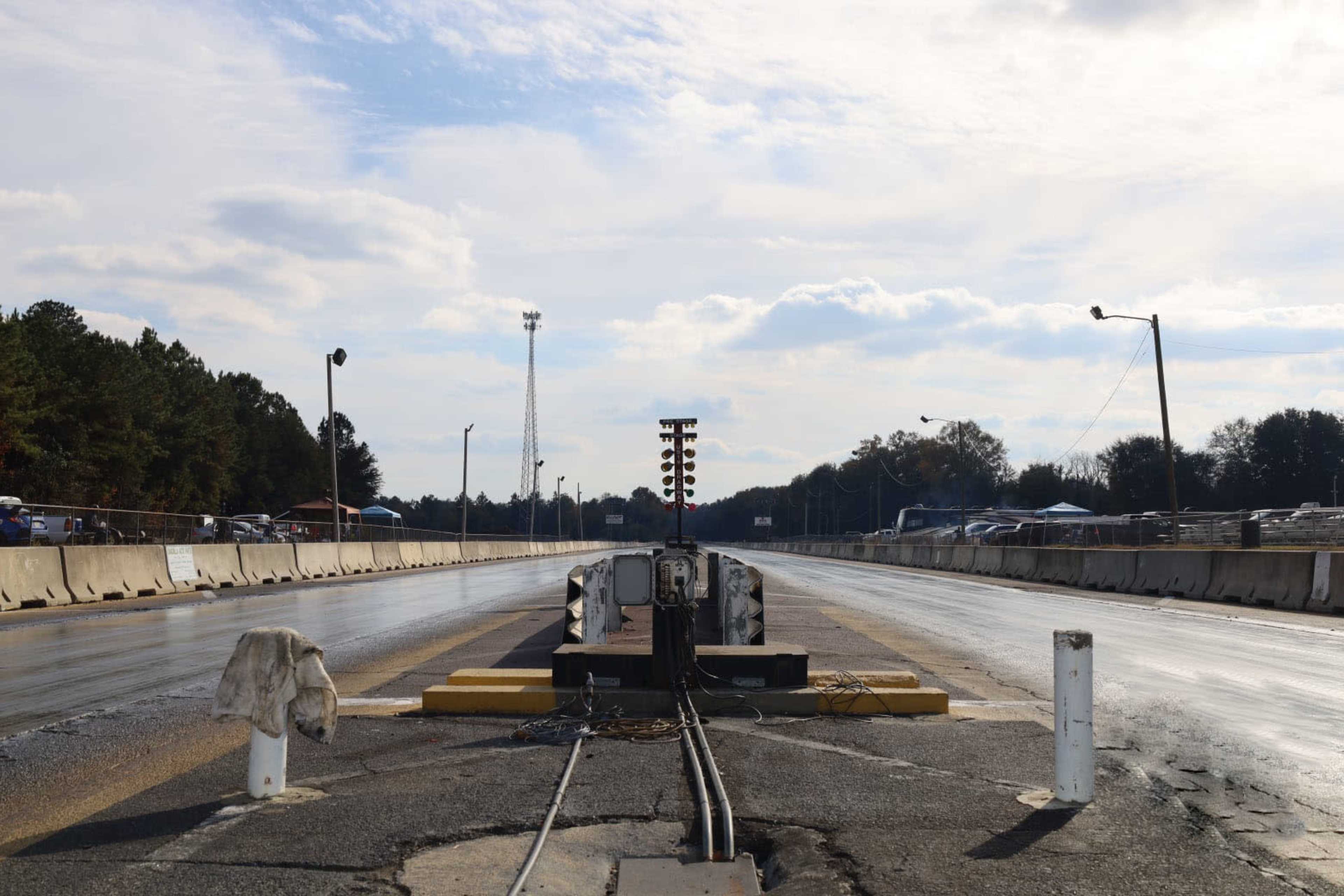 The image shows a drag racing track with a starting light system at the end, bordered by concrete barriers and trees in the background under a cloudy sky.