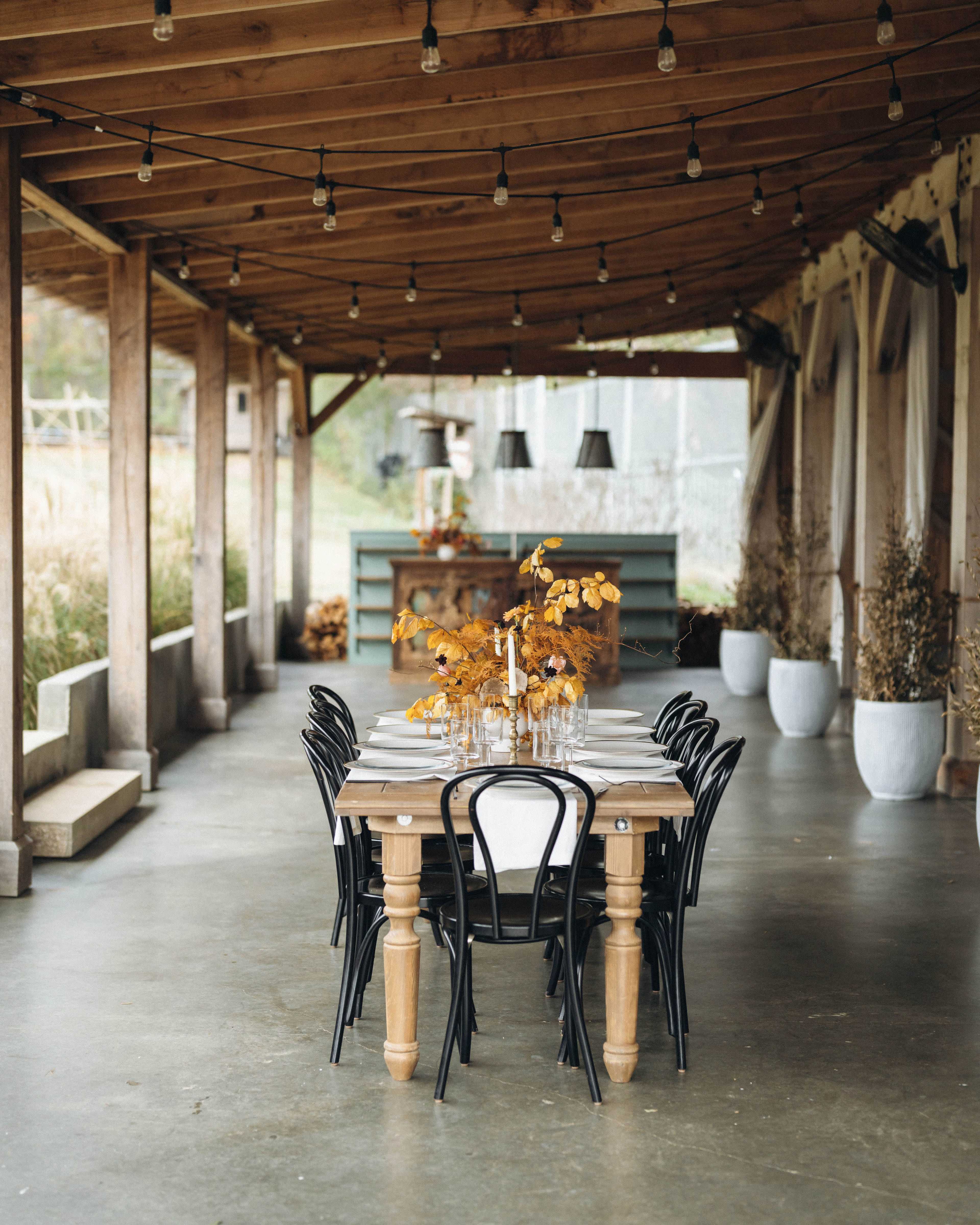 A long wooden dining table is set in a covered outdoor space, surrounded by black chairs and decorated with a floral centerpiece under string lights.