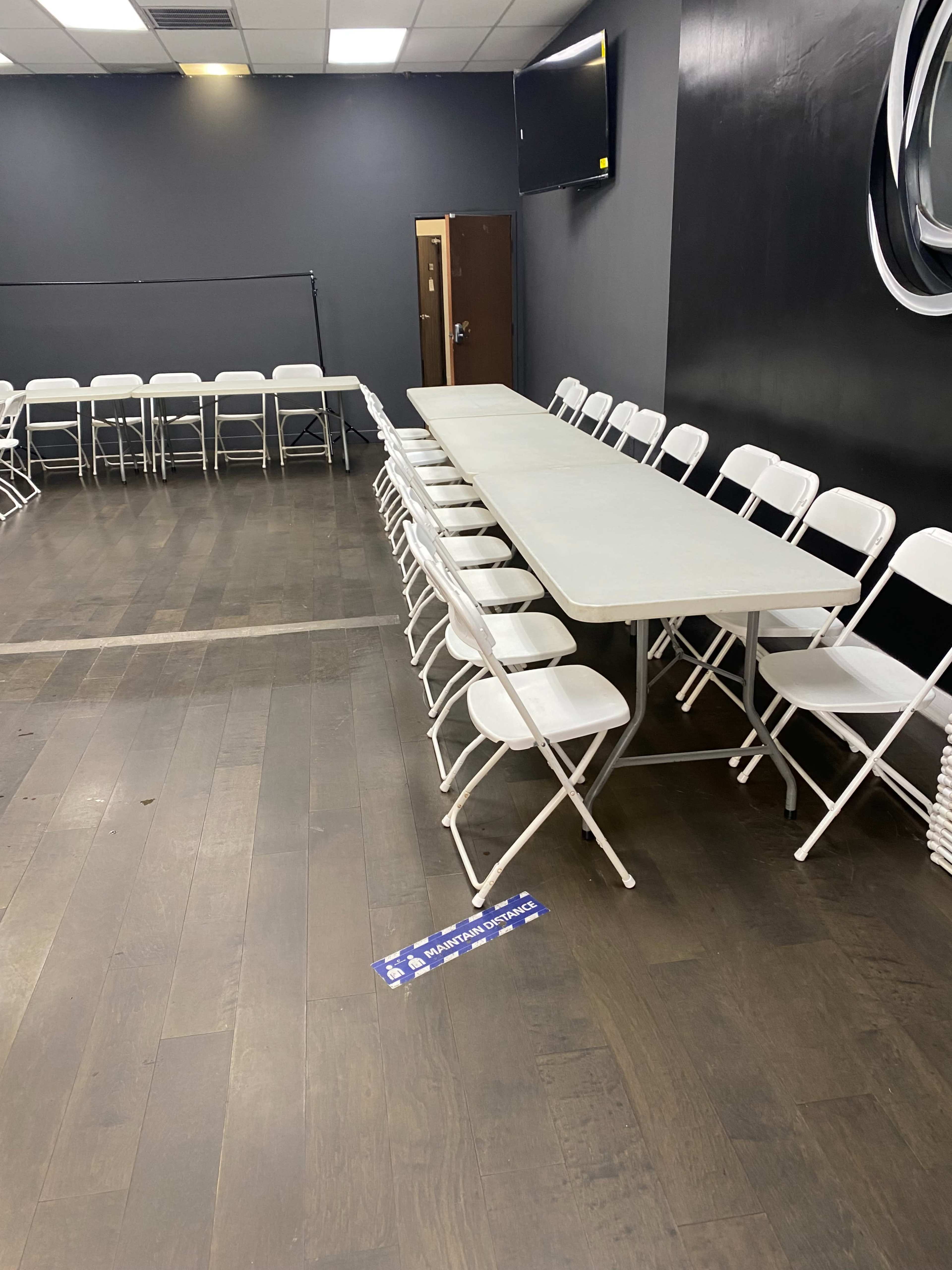 A long light-colored table is flanked by white folding chairs in a room with dark walls and a television mounted on one side.